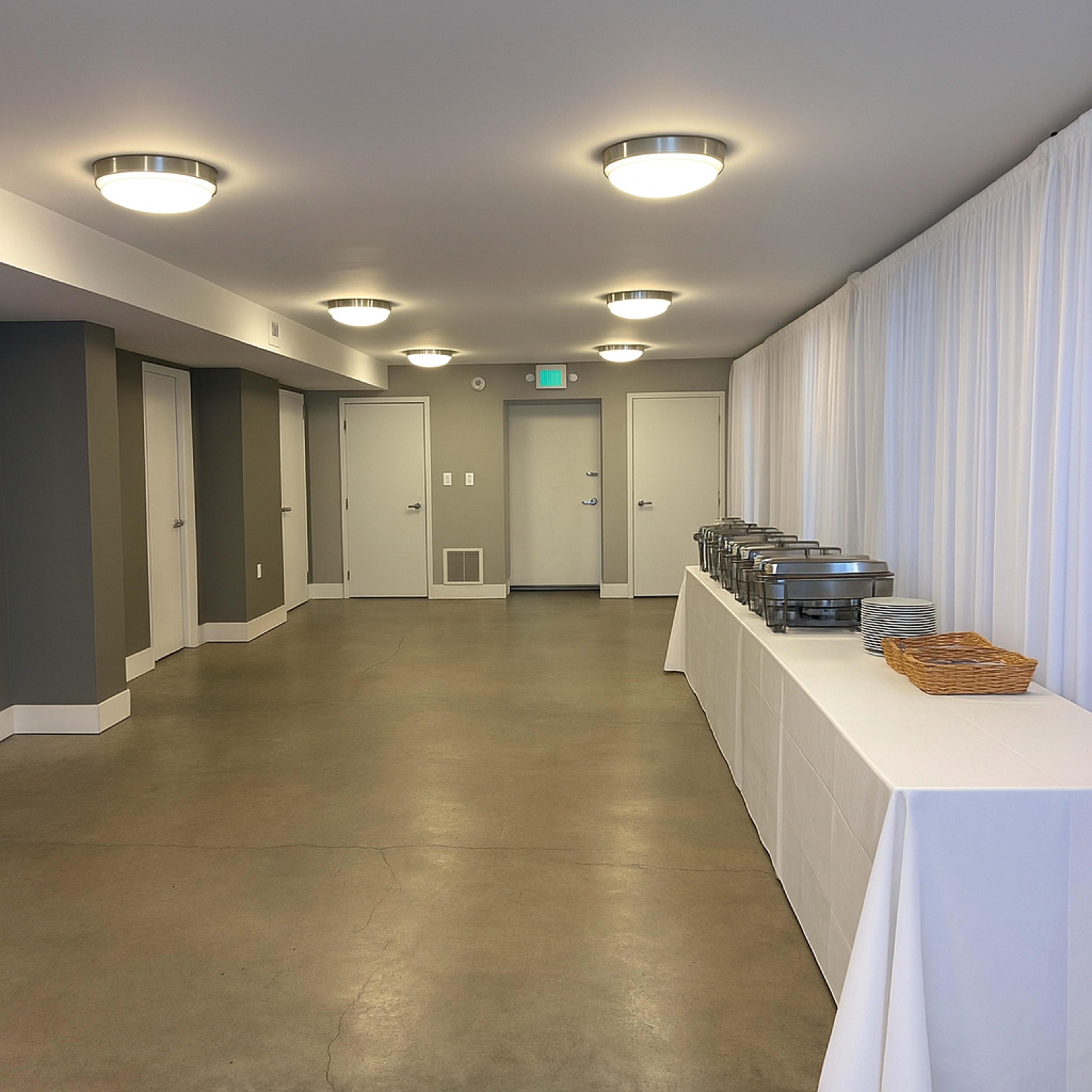 The image shows a modern, neutral-toned hallway with several closed doors and a long table covered by a white tablecloth displaying food trays and a basket.