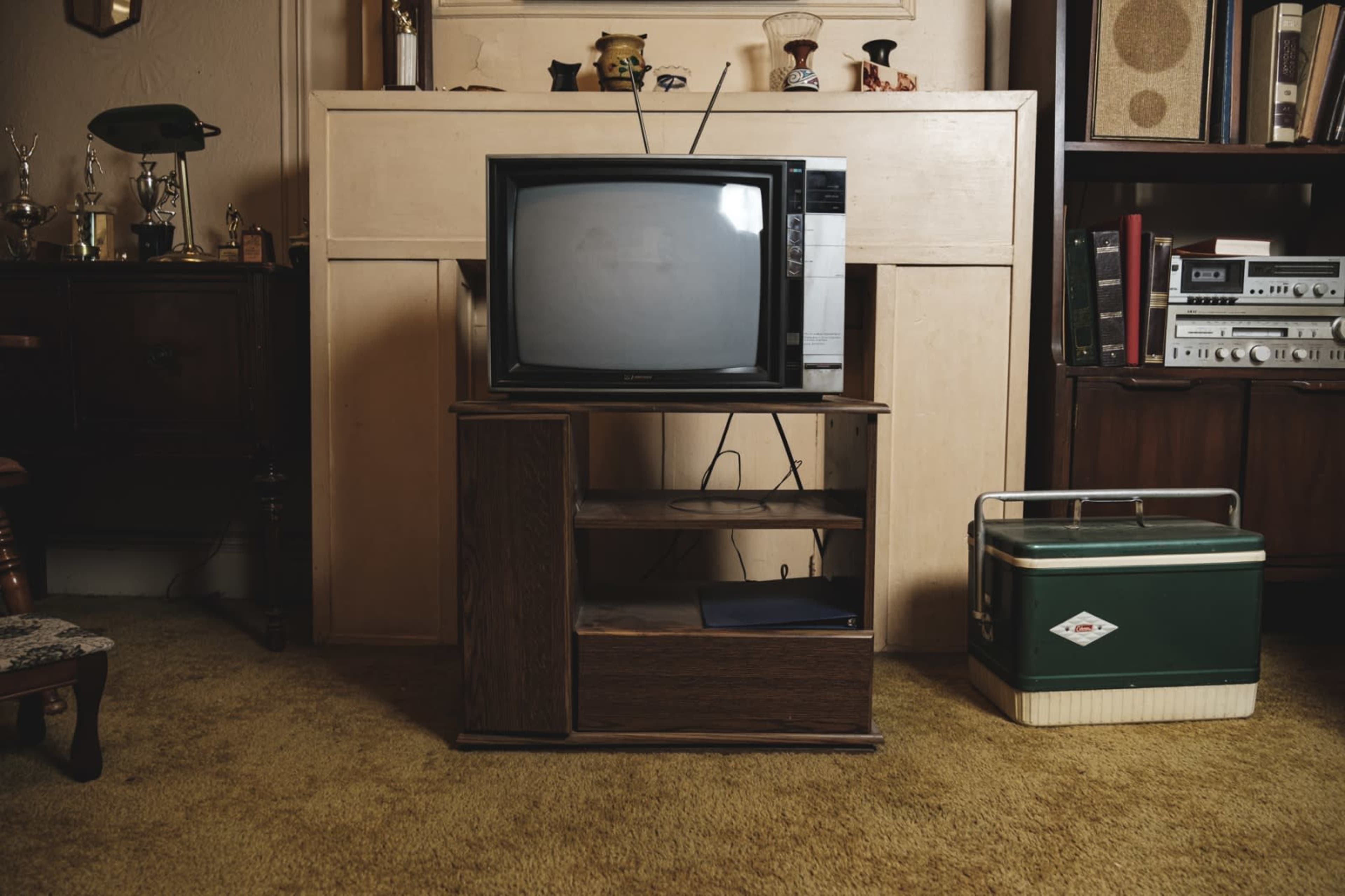 A retro living room features an old television on a wooden stand, set against a beige mantel, with a green cooler placed nearby.