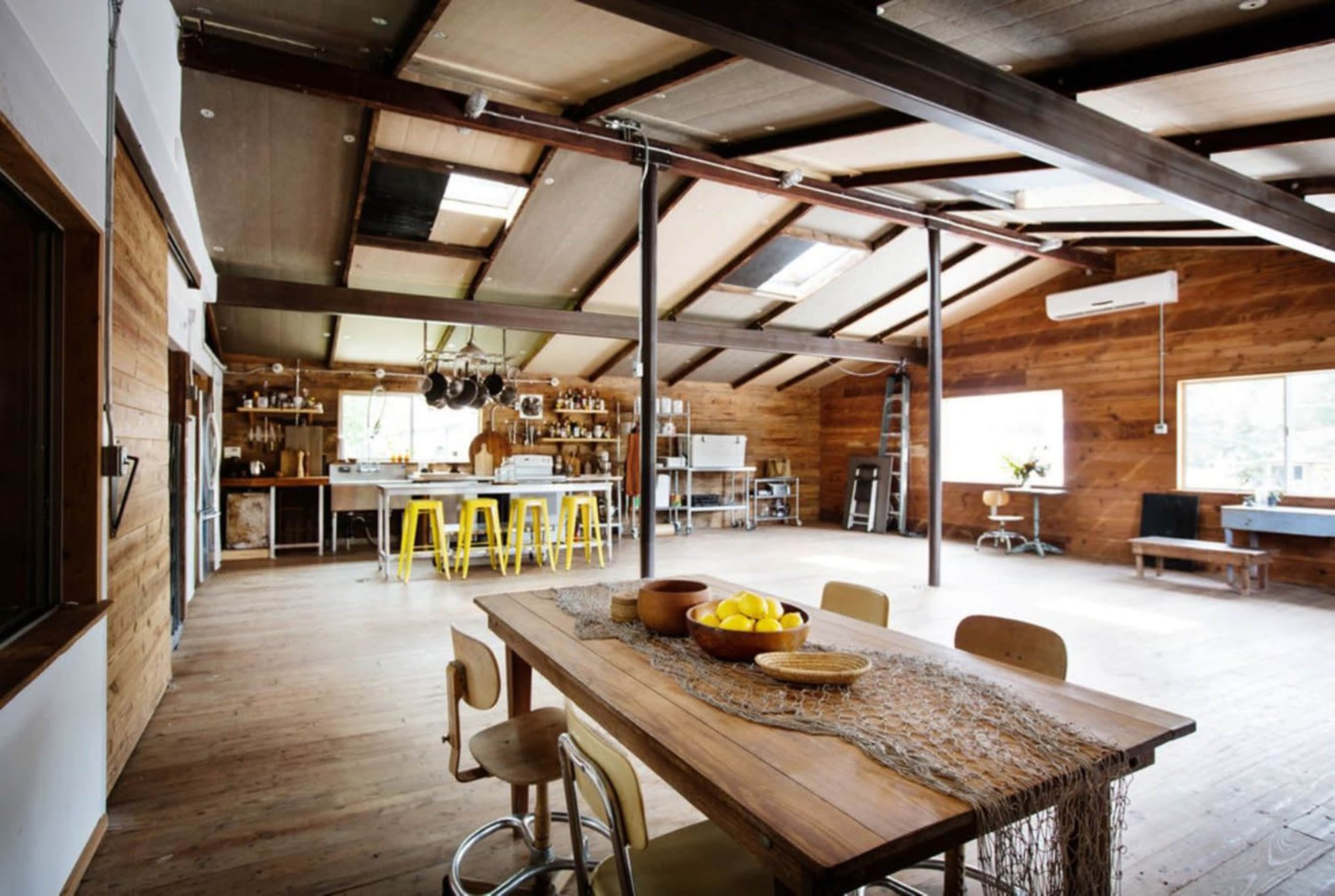A spacious, modern kitchen and dining area with wooden walls, a large table, yellow stools, and an organized kitchen in the background.