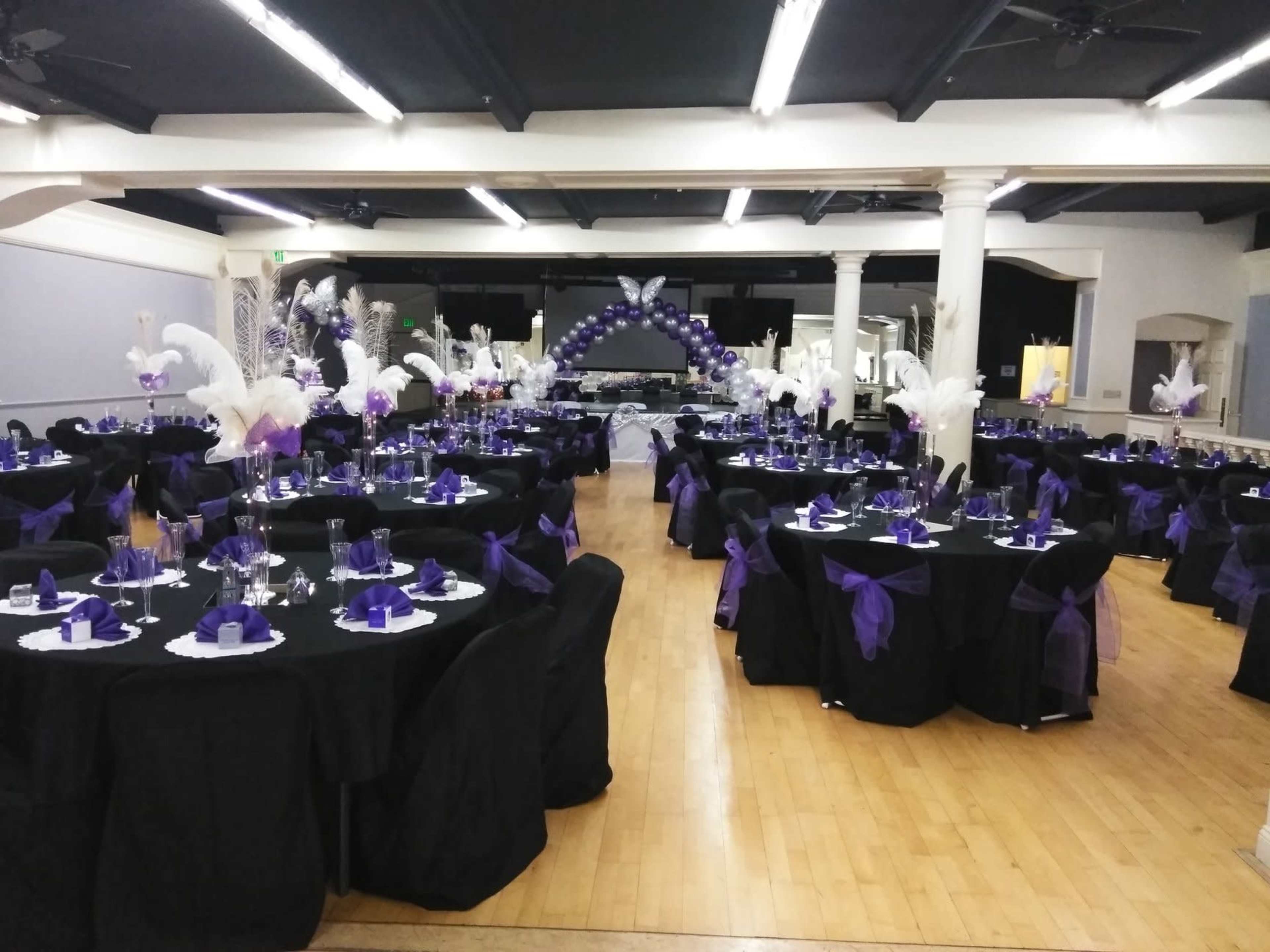 The image shows a banquet hall decorated for an event, featuring black tables with purple table settings and centerpieces, along with purple and white balloons and feather arrangements.