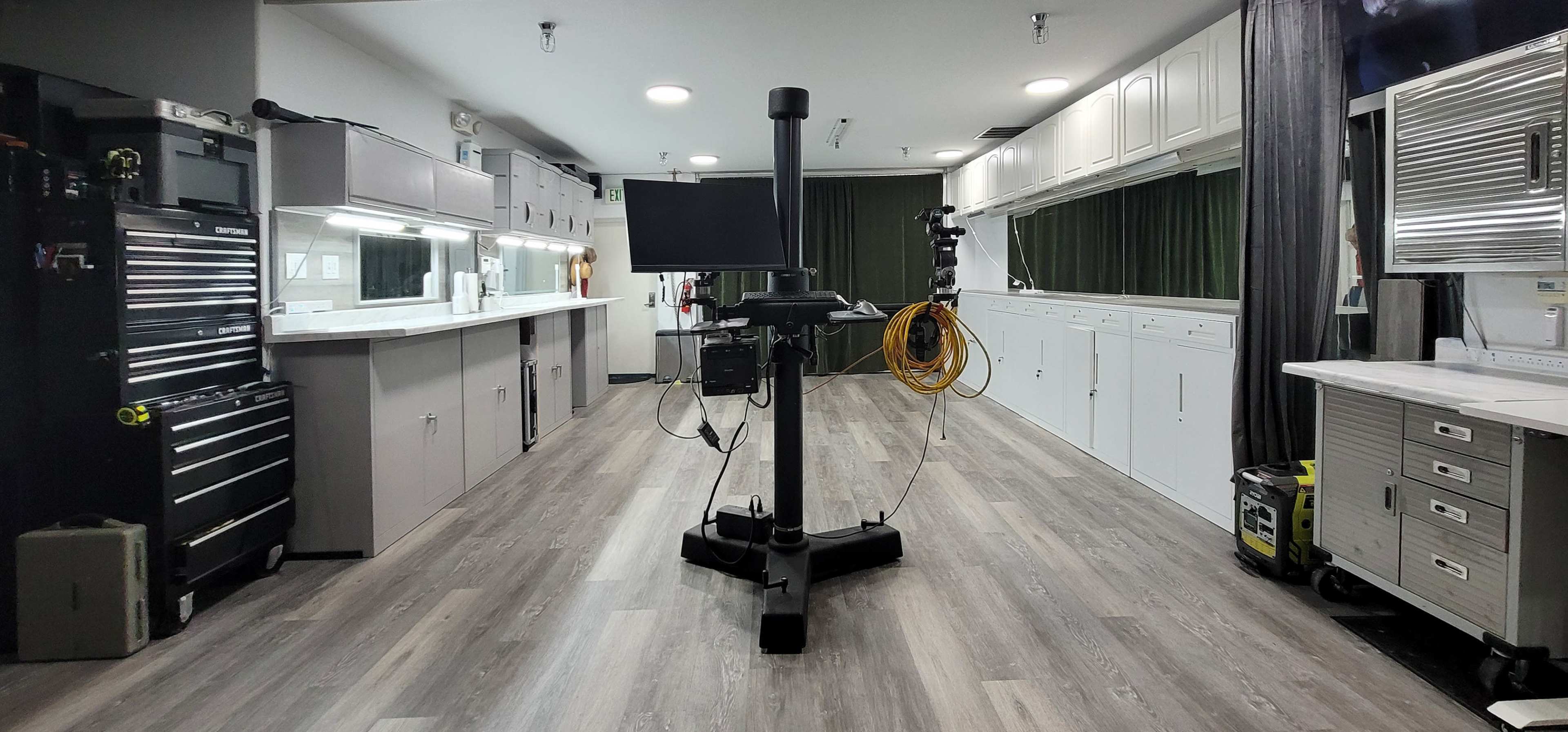 The image shows a room with a central black vertical stand equipped with a monitor, surrounded by cabinets and workstations on either side, all set on a wooden floor.