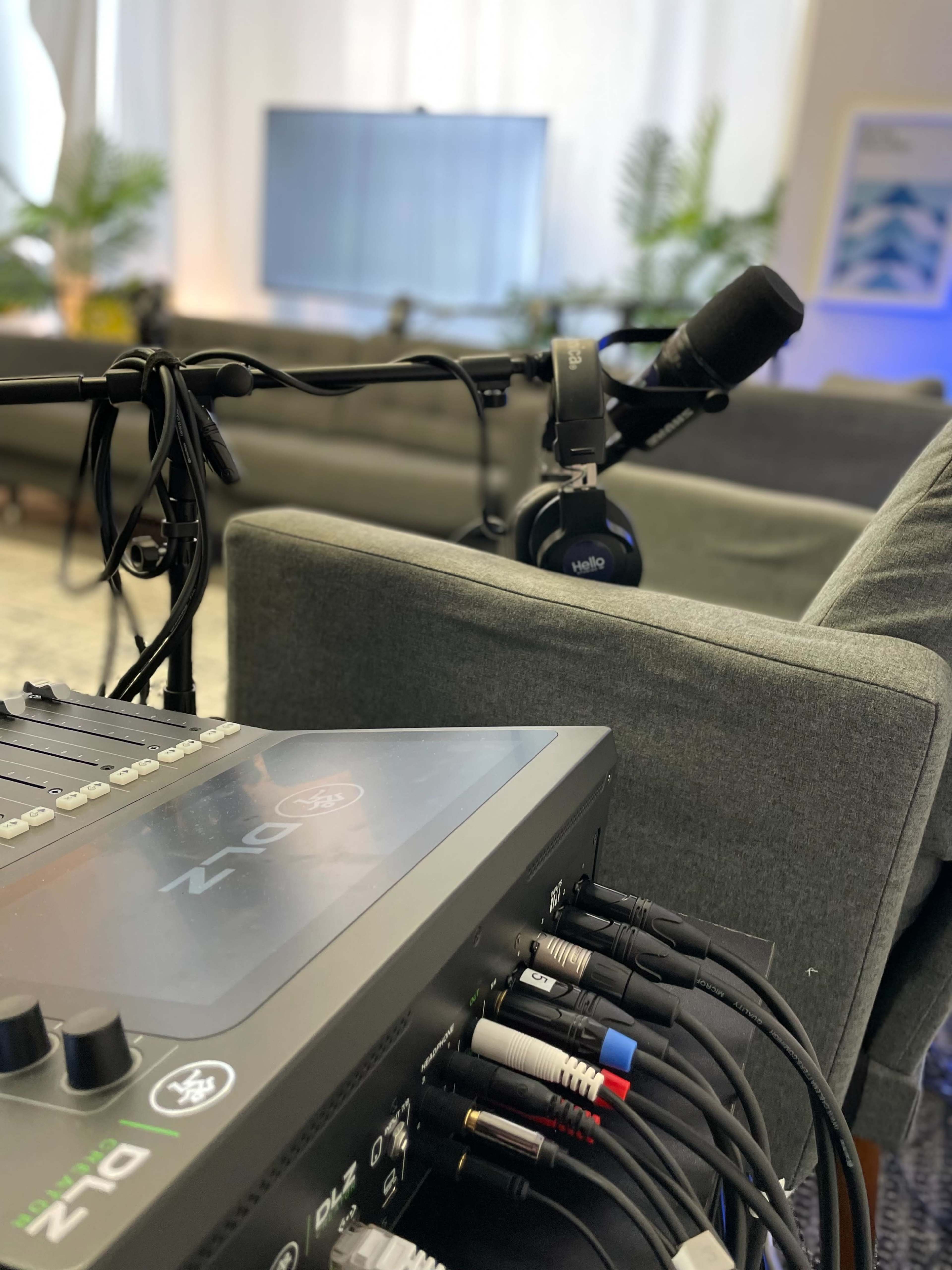 A mixing console is positioned next to a microphone and headphones, with a blurred living room in the background.