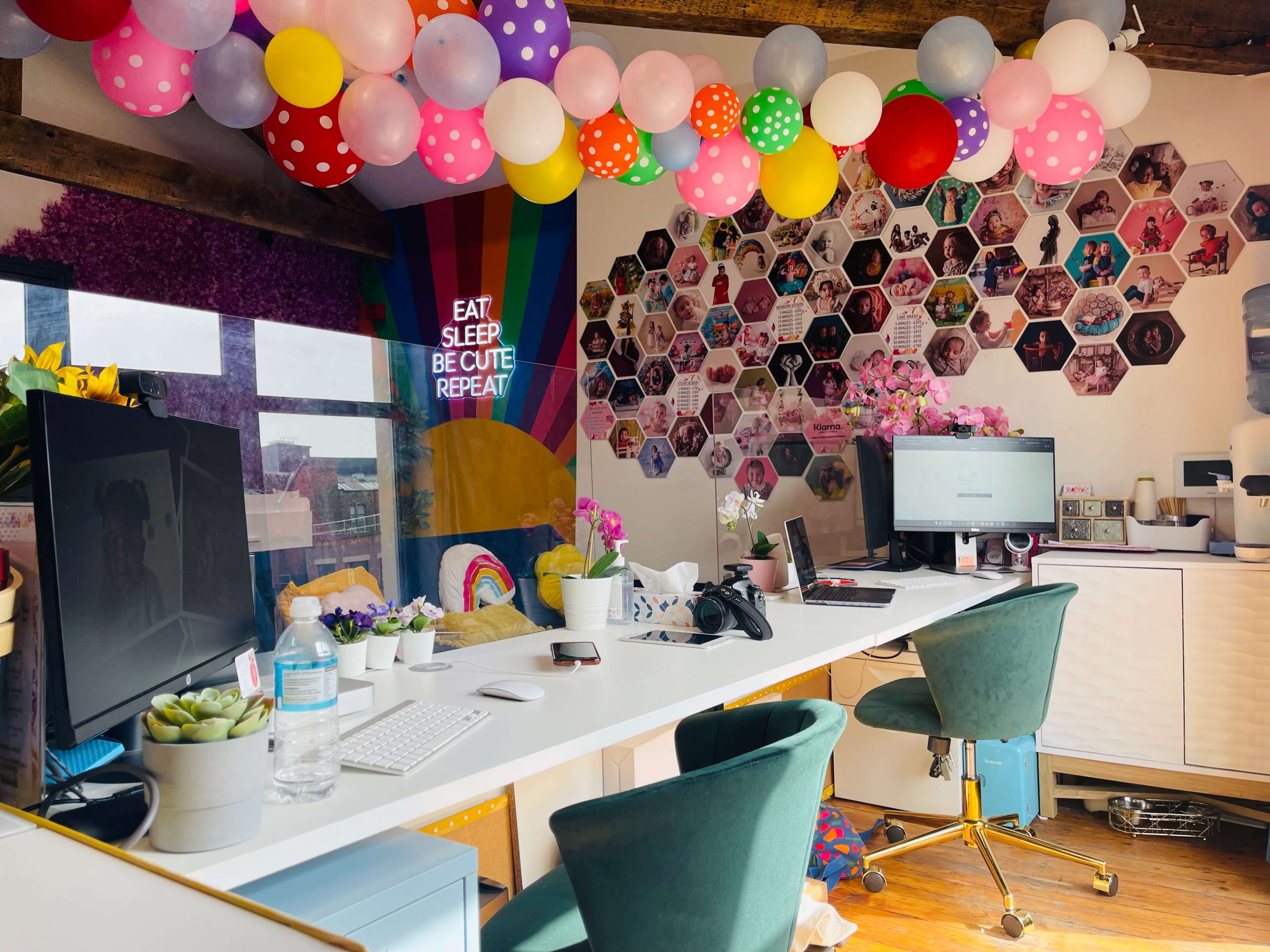 A colorful office space decorated with balloons, personal photos on the wall, and two desks equipped with computers and plants.