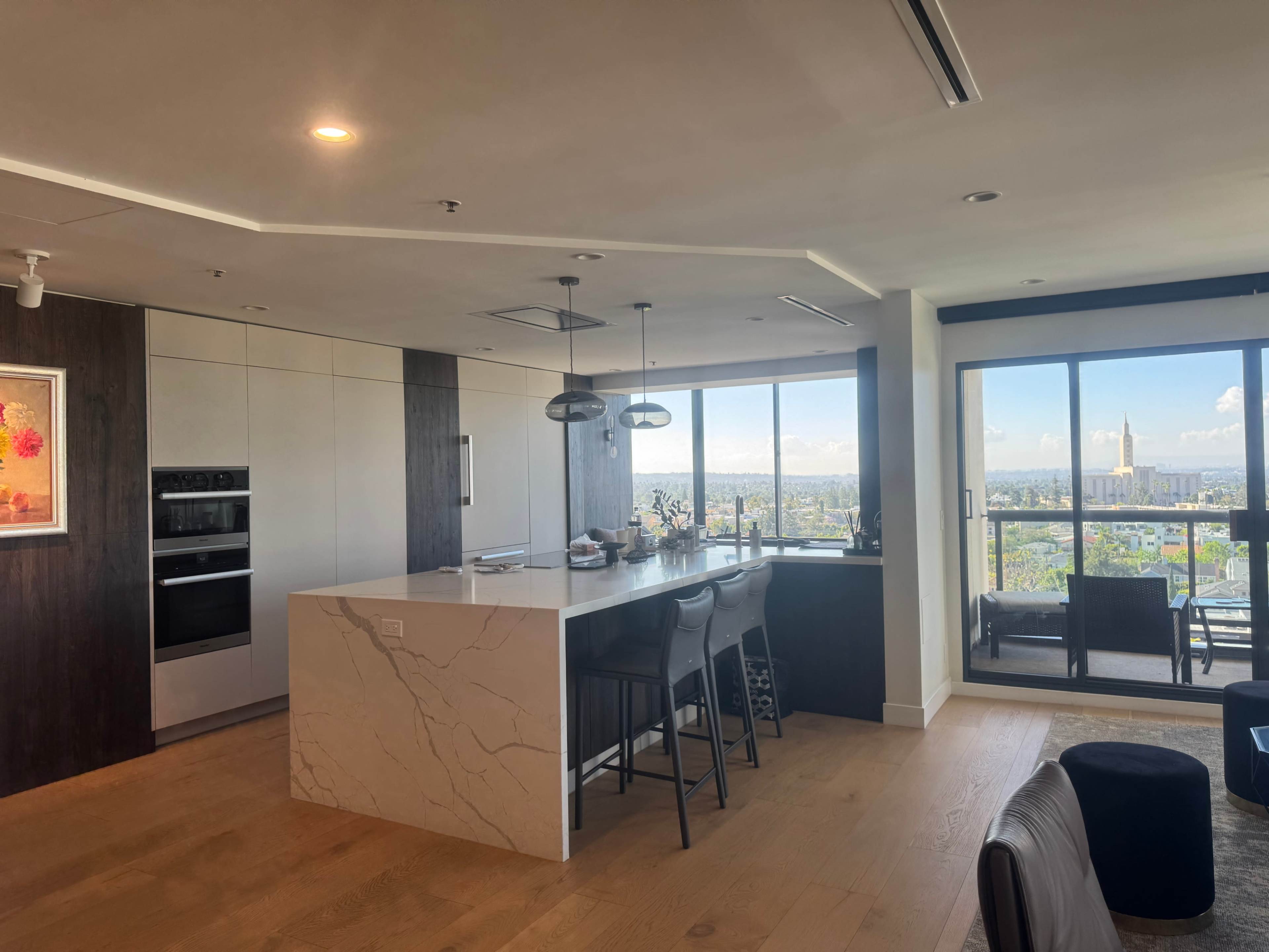 The image shows a modern kitchen and dining area with a large island, sleek cabinetry, and a panoramic view of a city skyline through a glass balcony door.