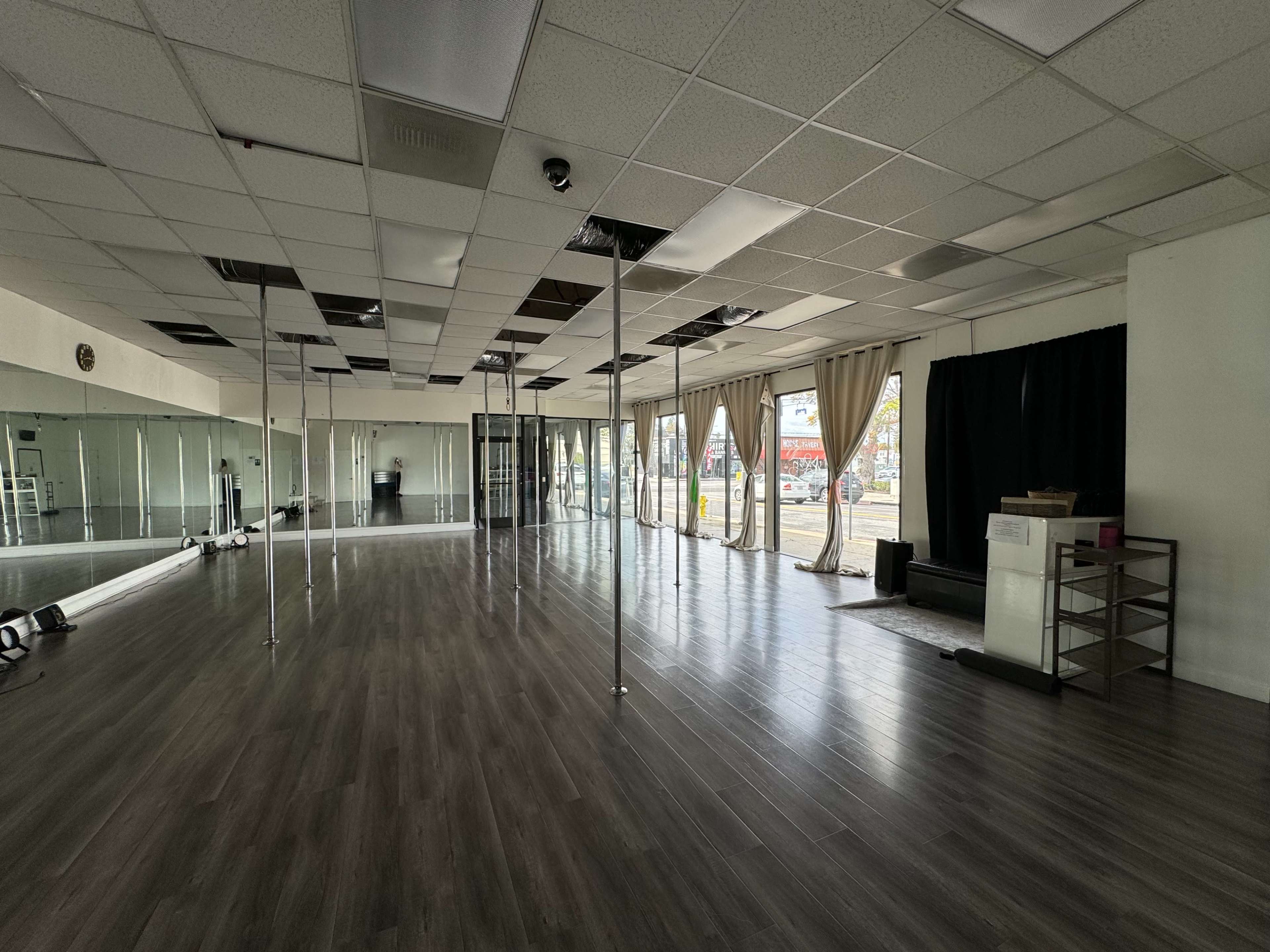 An empty dance studio with several dance poles, a mirrored wall, and large windows revealing an outdoor view.