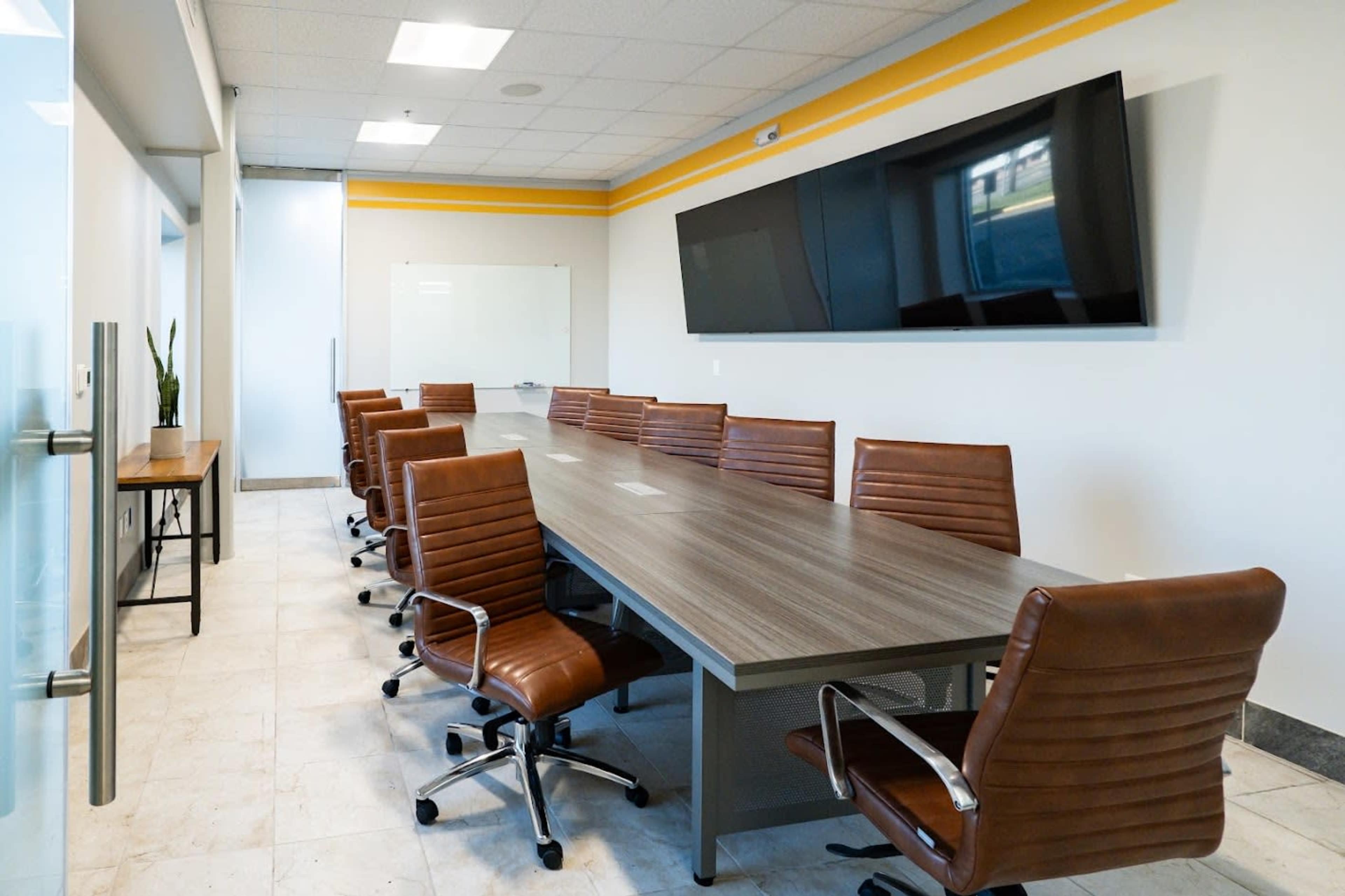 A long wooden conference table is surrounded by brown leather chairs in a well-lit meeting room with a large screen mounted on the wall.