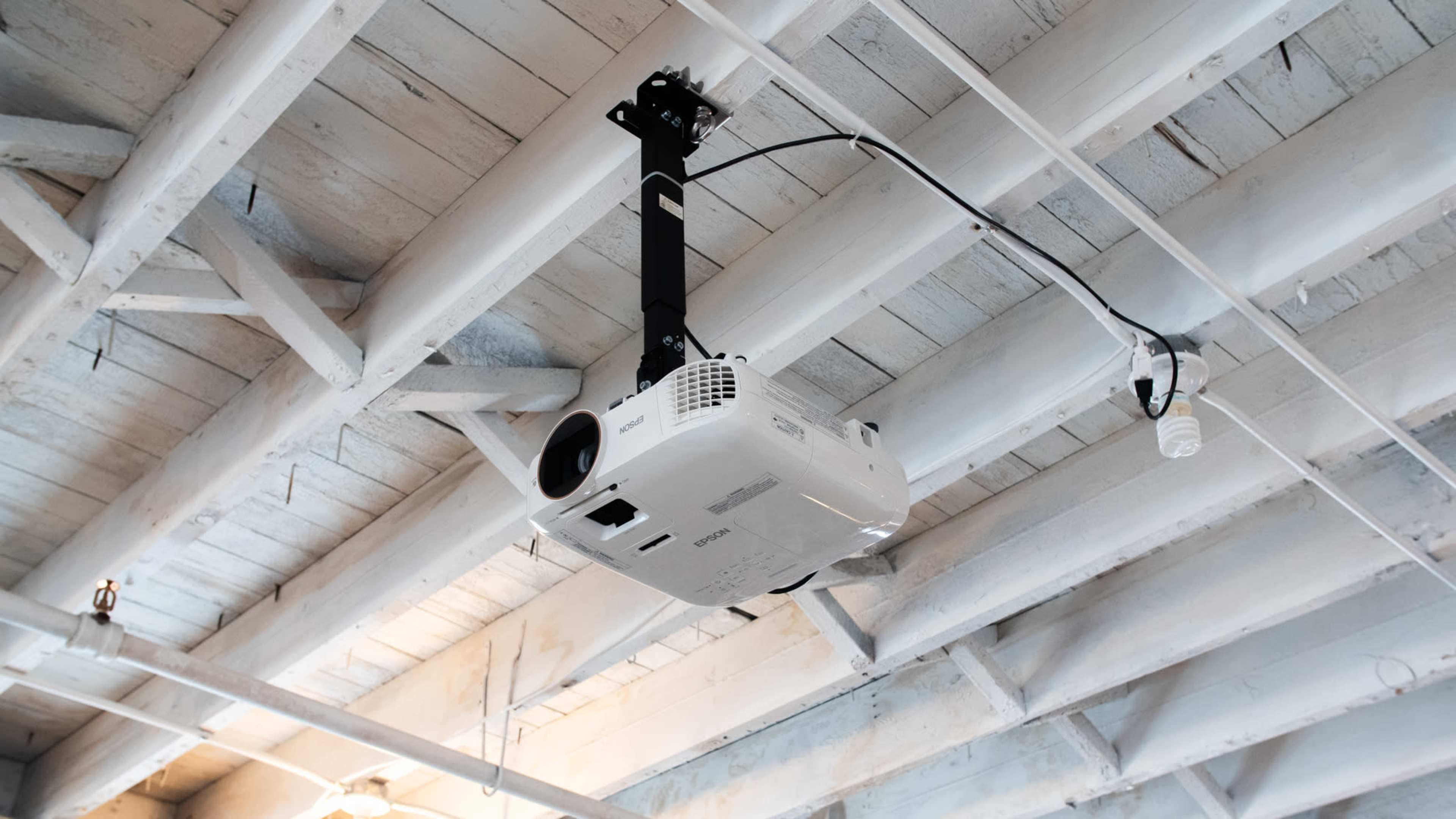 A ceiling-mounted projector is suspended from white wooden beams in a room.