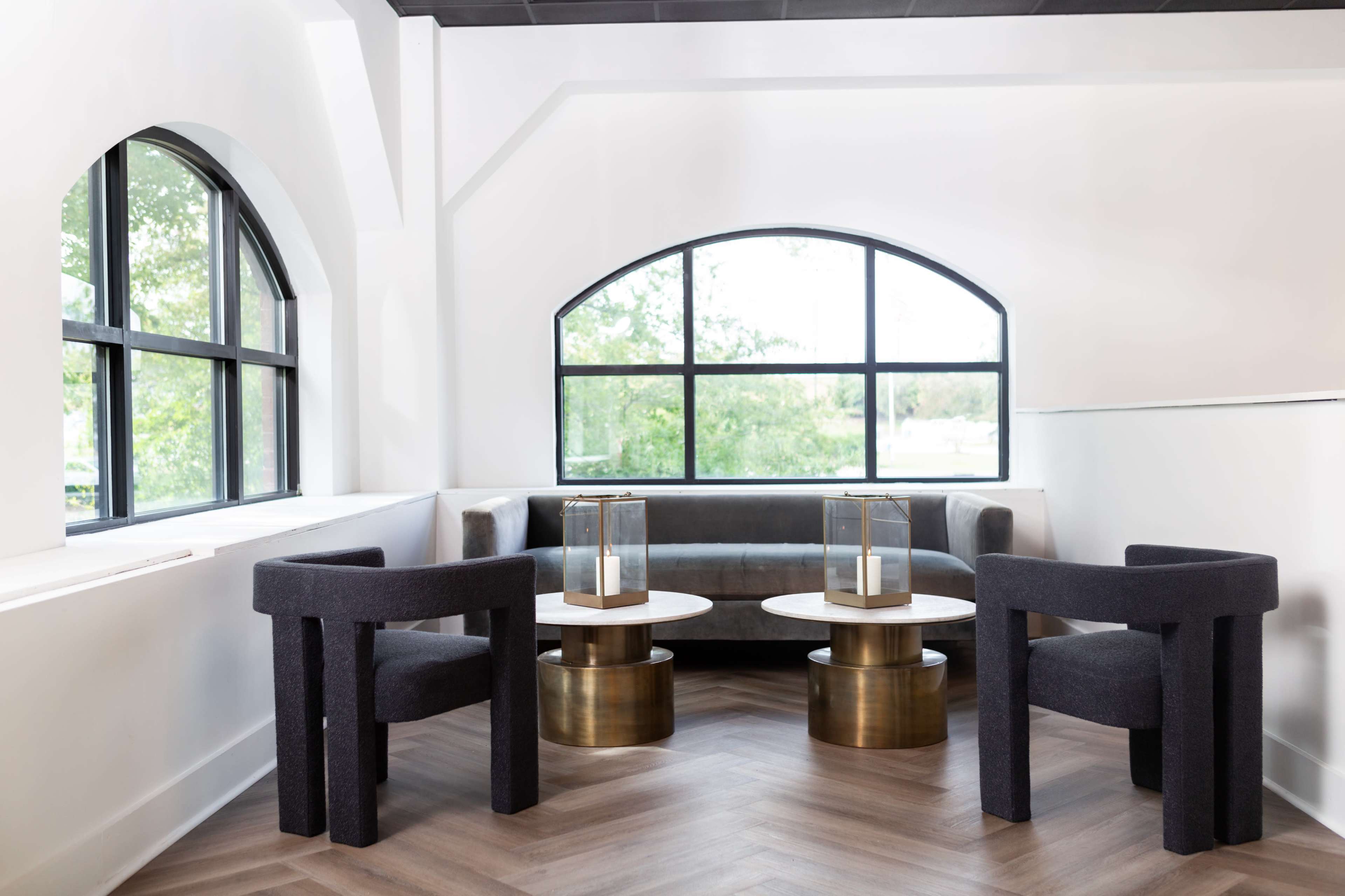 A modern seating area features two dark fabric chairs and two circular tables, one wood and one metallic, situated by large windows.