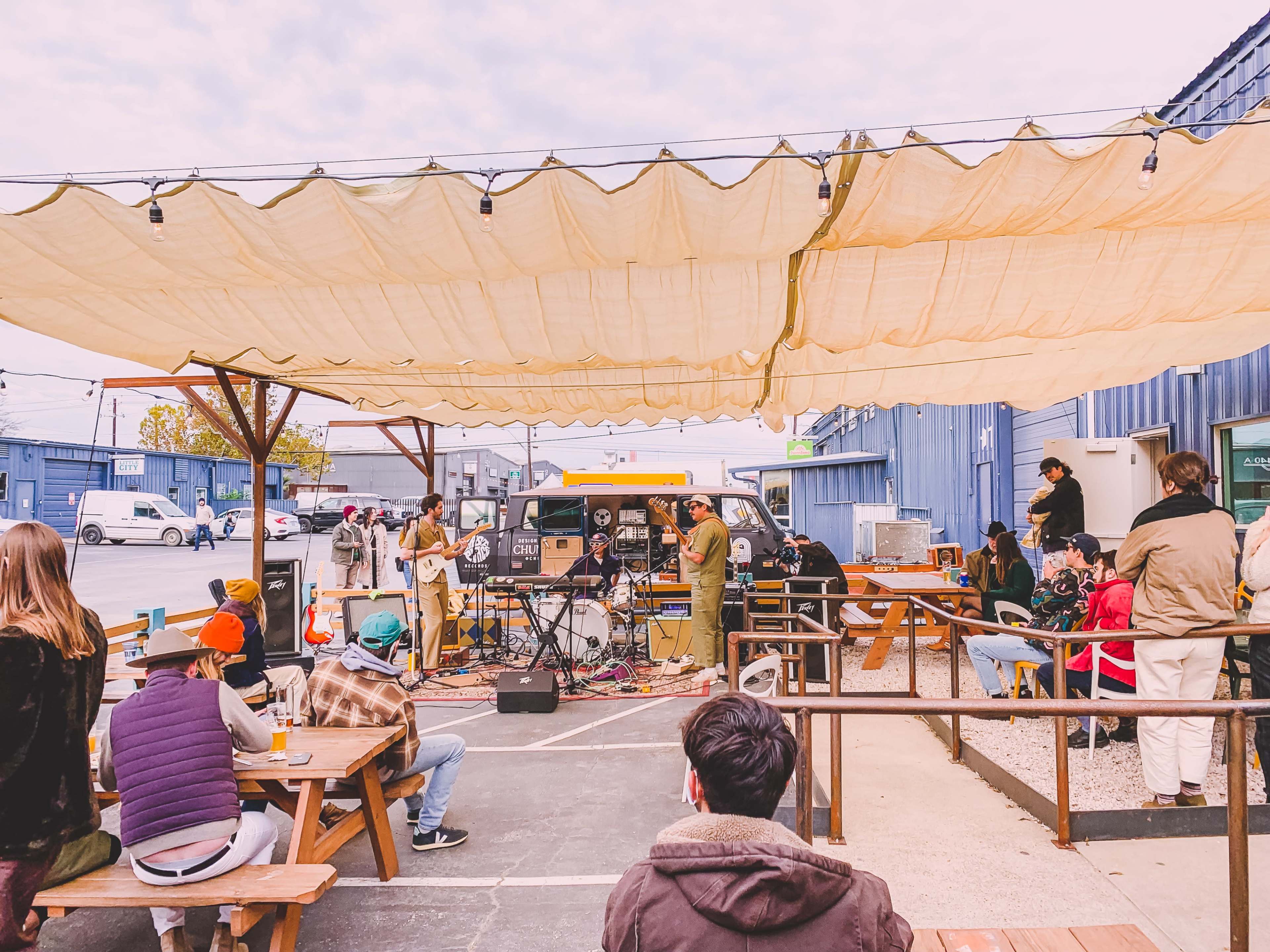 A band performs on a small stage under a large canopy while an audience sits at wooden tables outdoors.