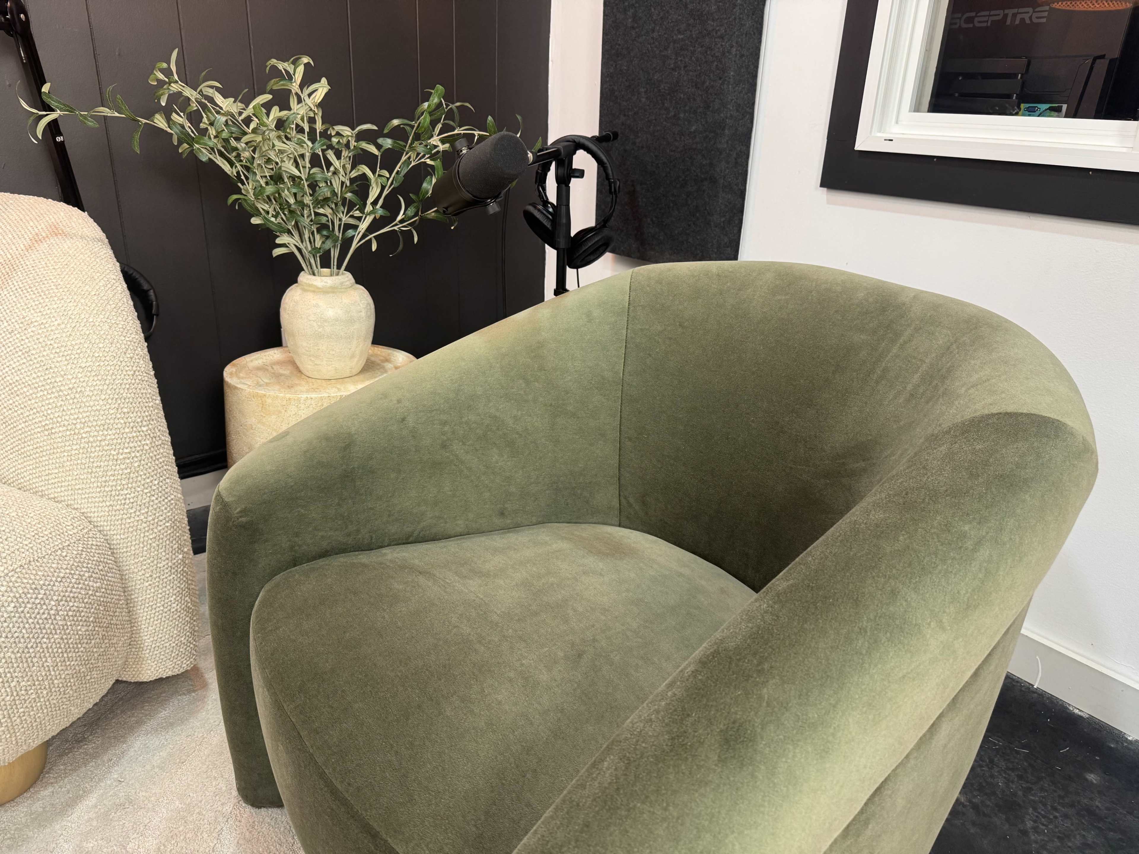 A curved, green velvet armchair is positioned beside a light-colored vase containing a plant, against a black and white backdrop.
