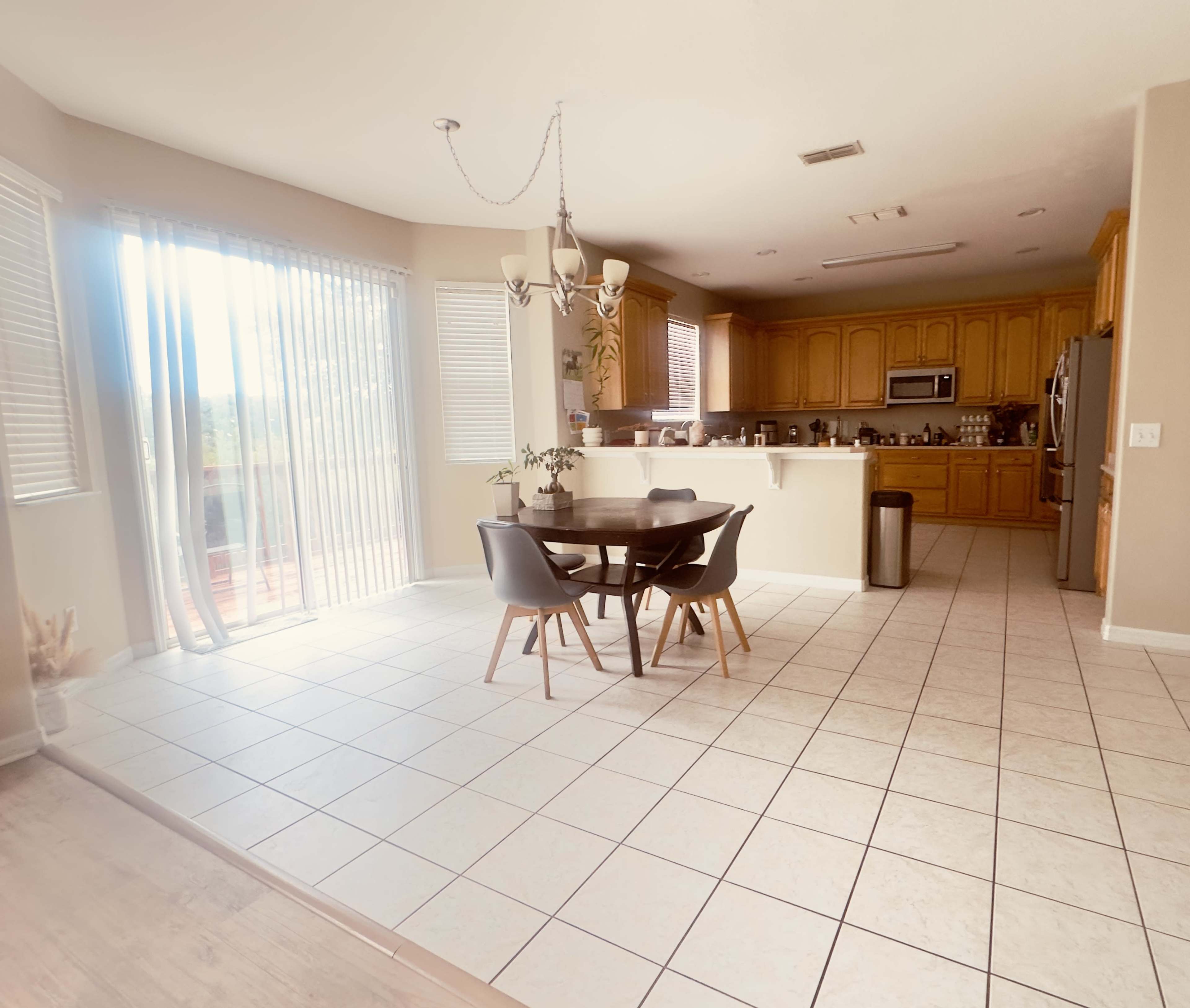 The image shows a spacious kitchen and dining area with a round table and chairs, large windows allowing natural light, and wooden cabinetry in the kitchen.