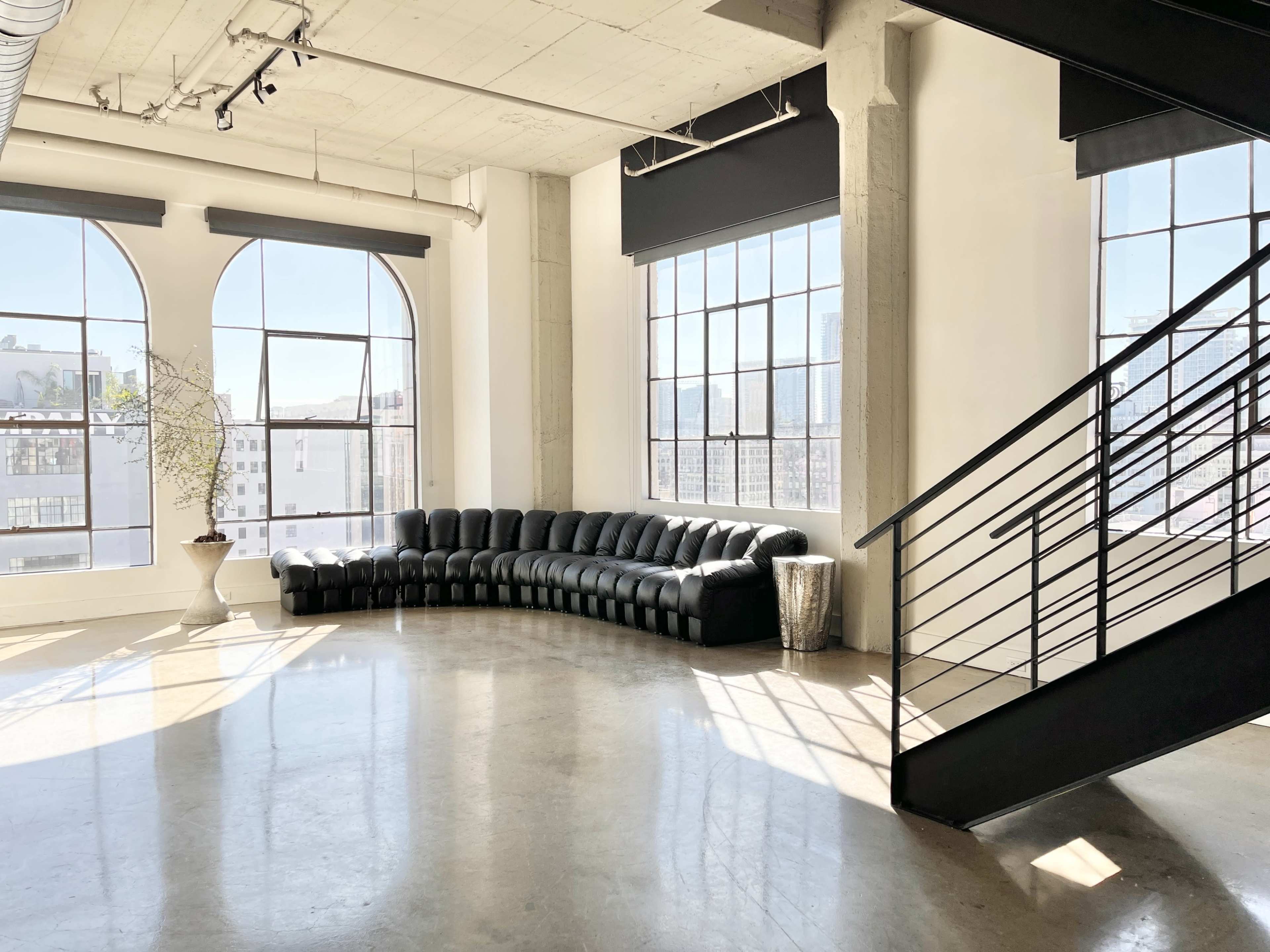 A curved black leather sofa is positioned along the wall beneath large windows in a bright, open space with concrete floors and a staircase nearby.