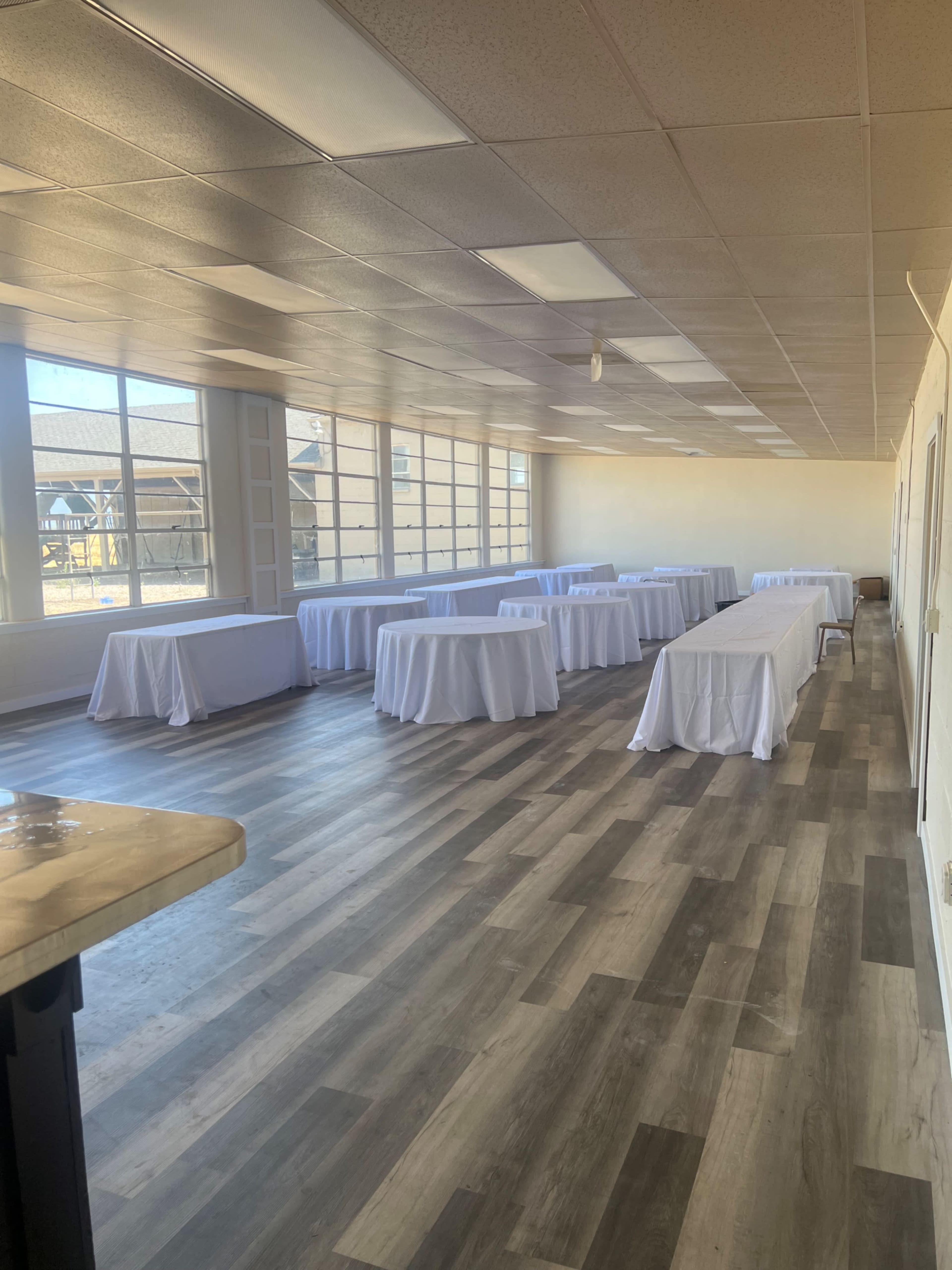 A spacious room with multiple tables covered in white tablecloths, illuminated by natural light from large windows.
