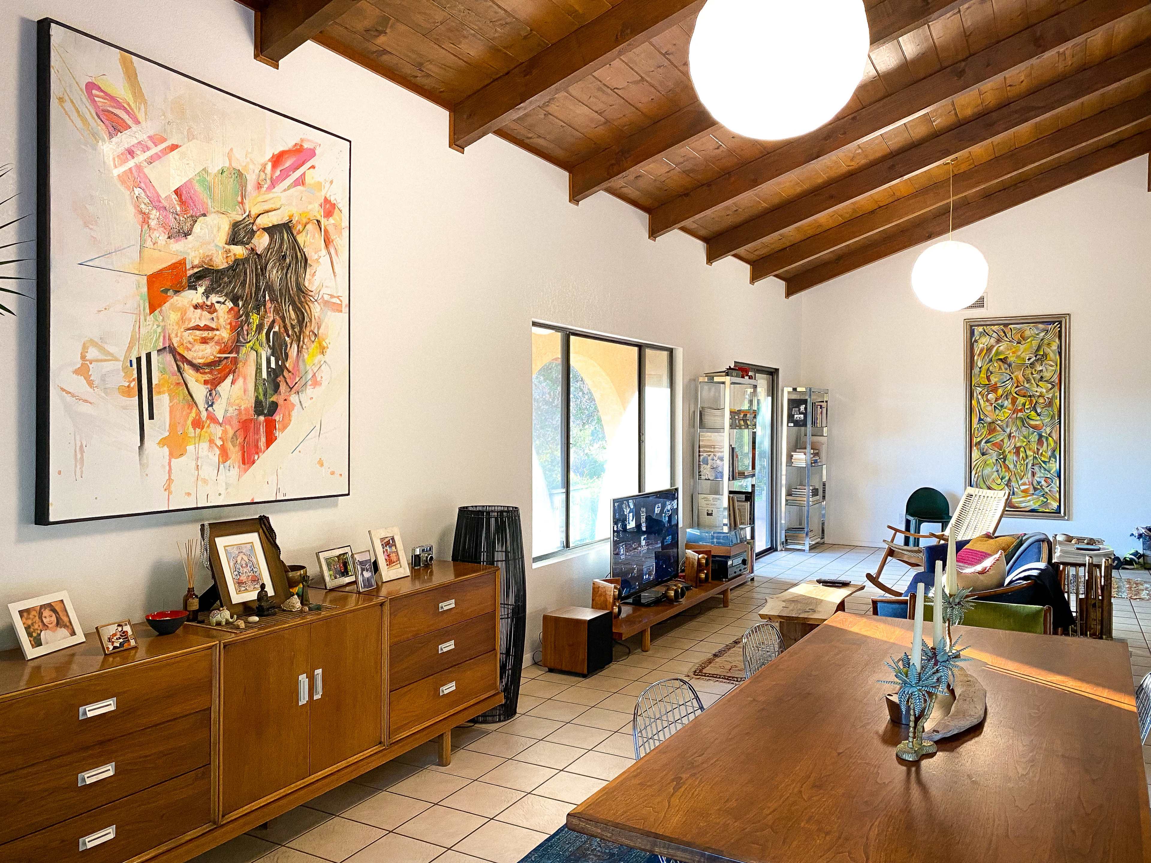 The image shows a spacious living area with a wooden ceiling, featuring a large colorful painting, a dining table, and various pieces of furniture.
