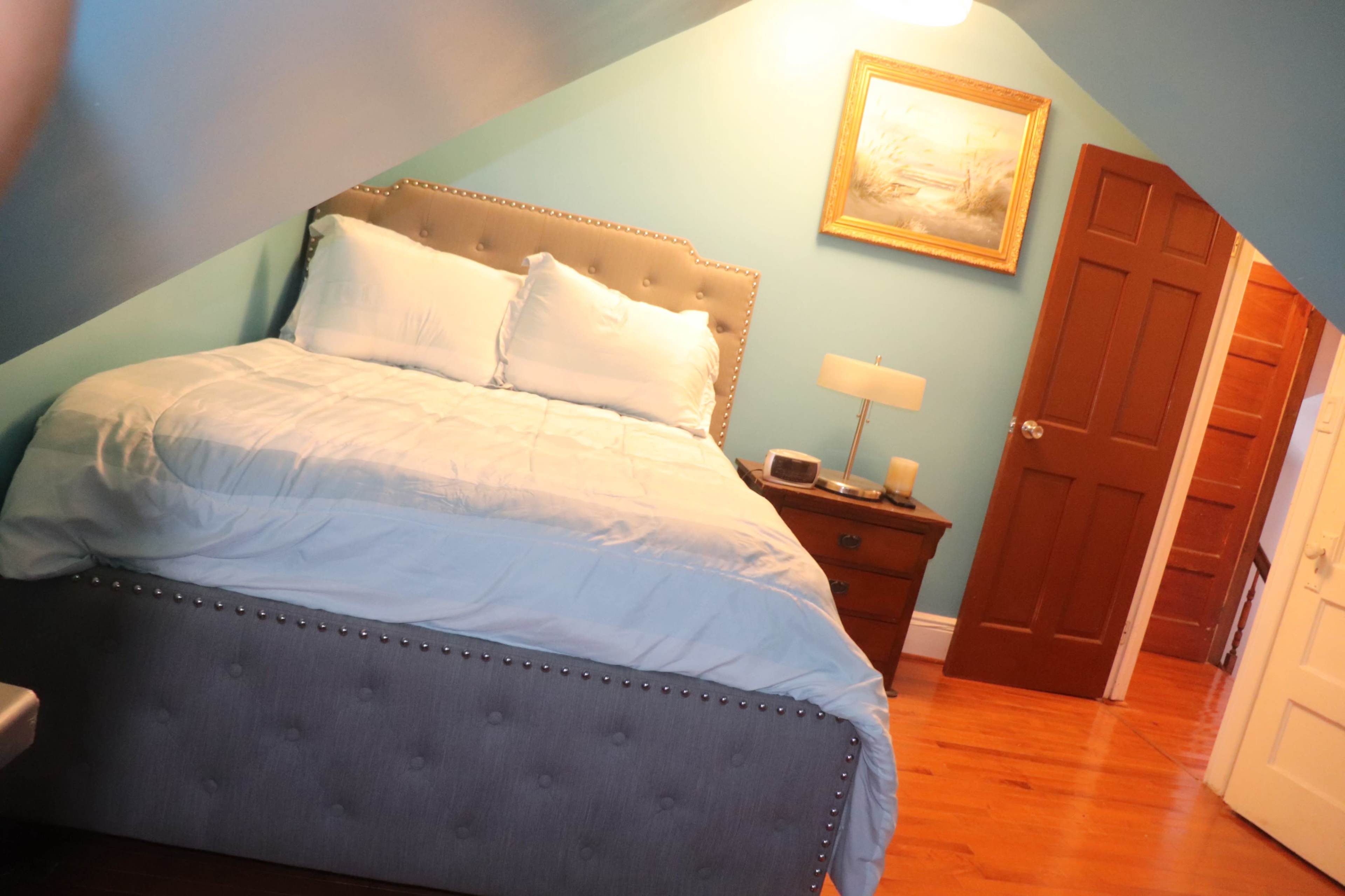 A bedroom with a bed, a nightstand, and a wooden door, featuring sloped ceilings and light blue walls.