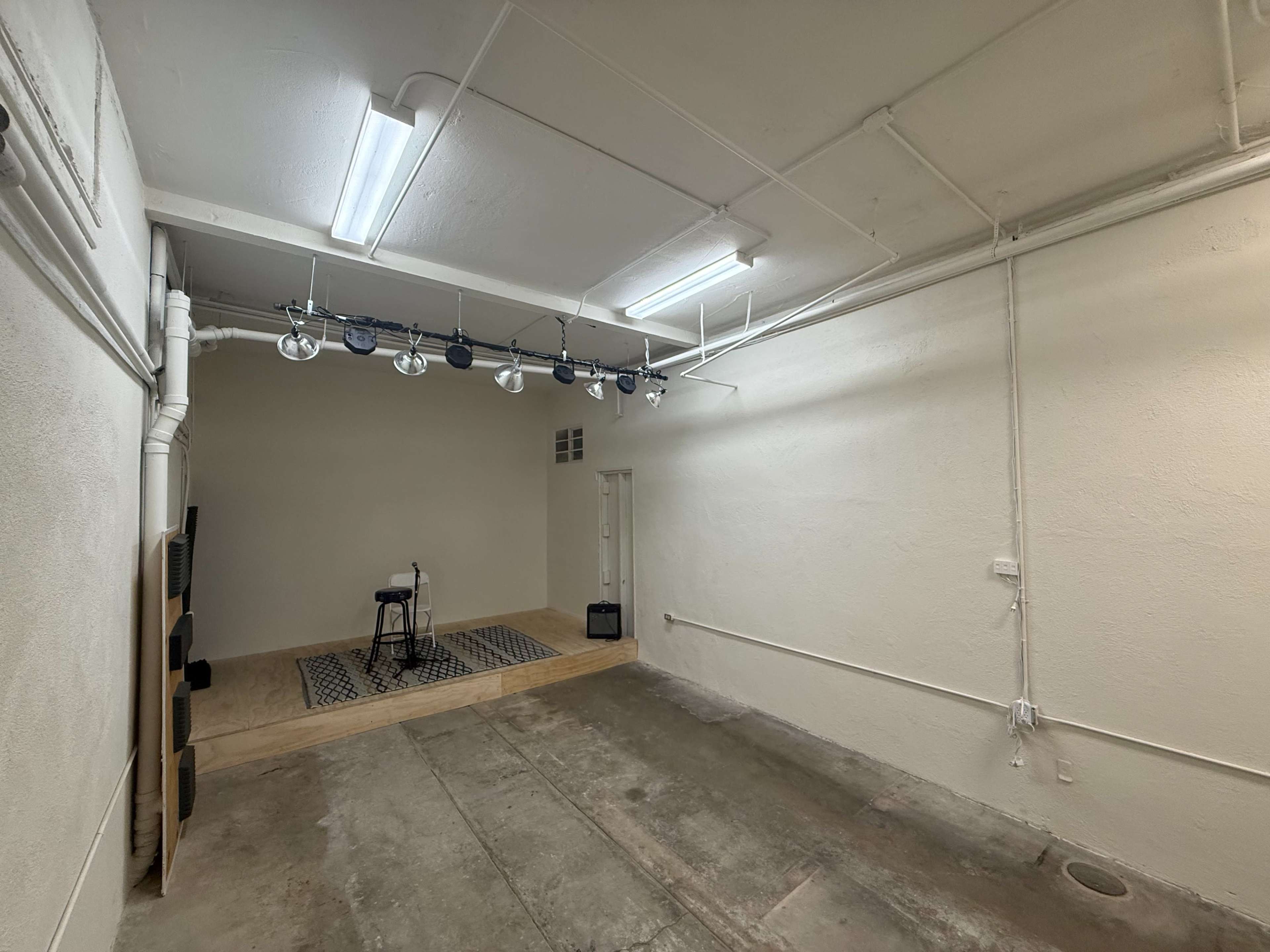 The image shows a spacious, empty room with concrete flooring, white walls, and a small stage area equipped with lighting and a microphone stand.