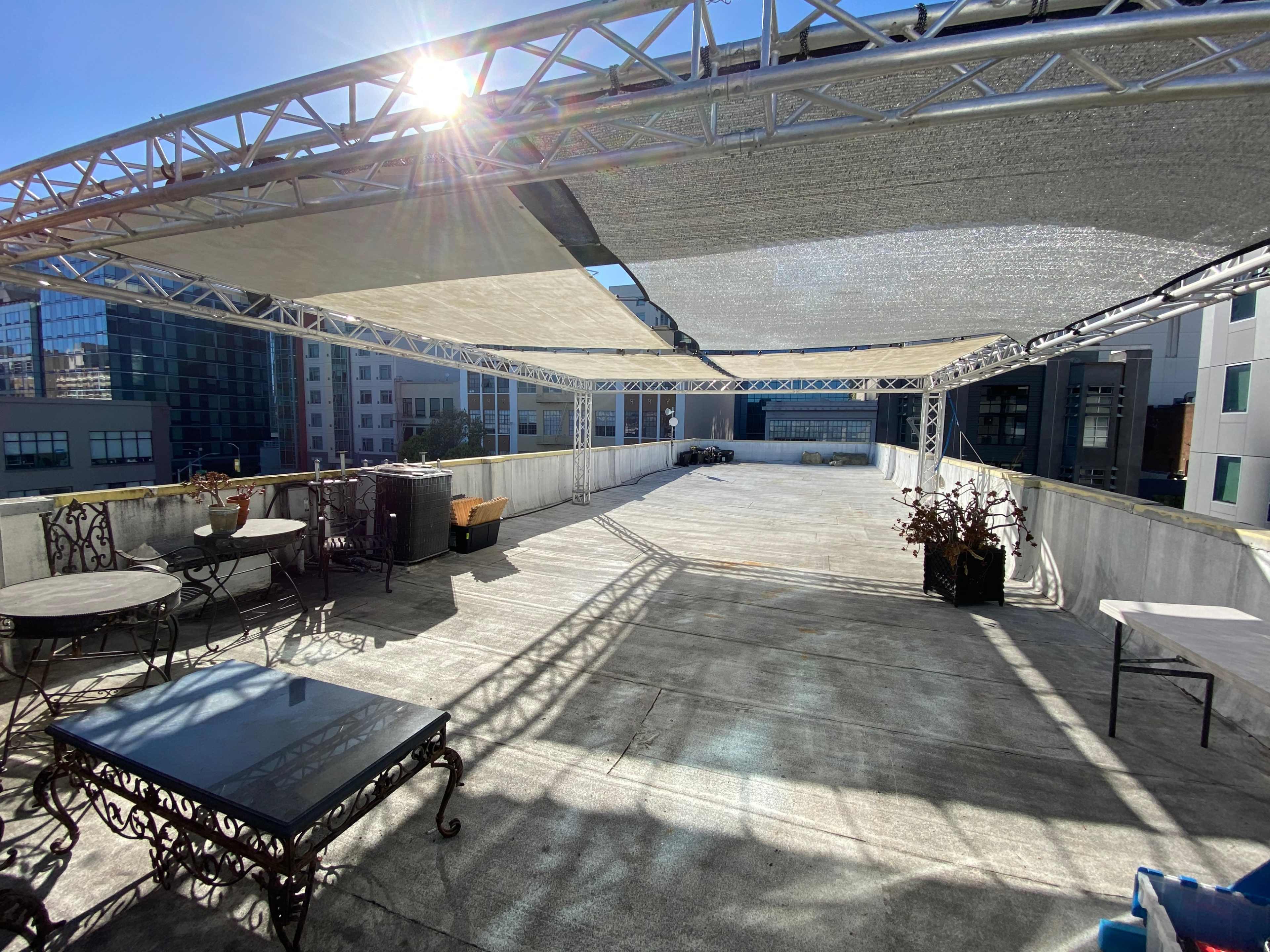 A rooftop space features a concrete floor with metal and glass furniture, shaded by fabric canopies, and surrounded by urban buildings.