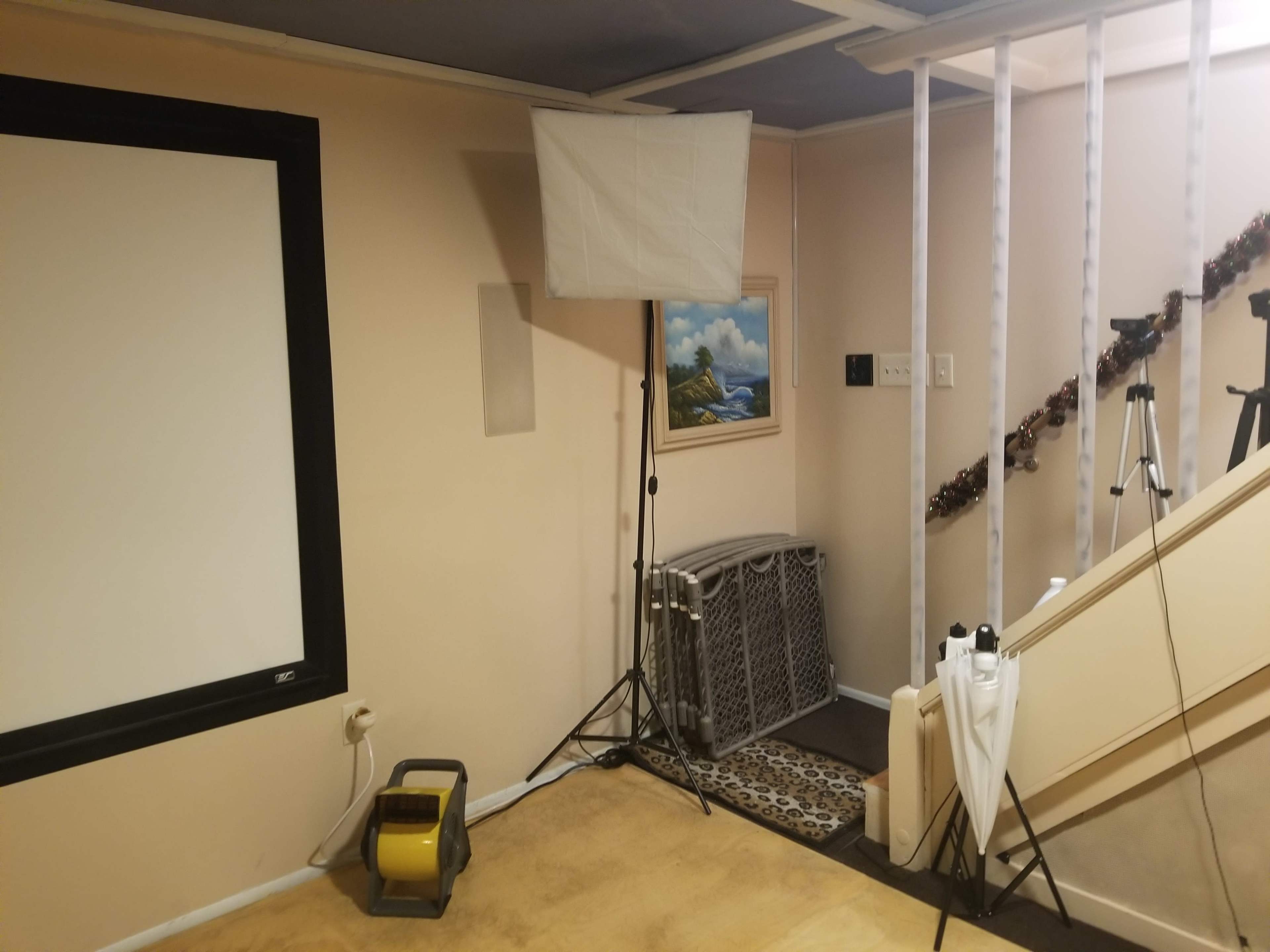 A basement room with a large white screen on one wall, a lighting setup on a tripod, and a staircase leading up.