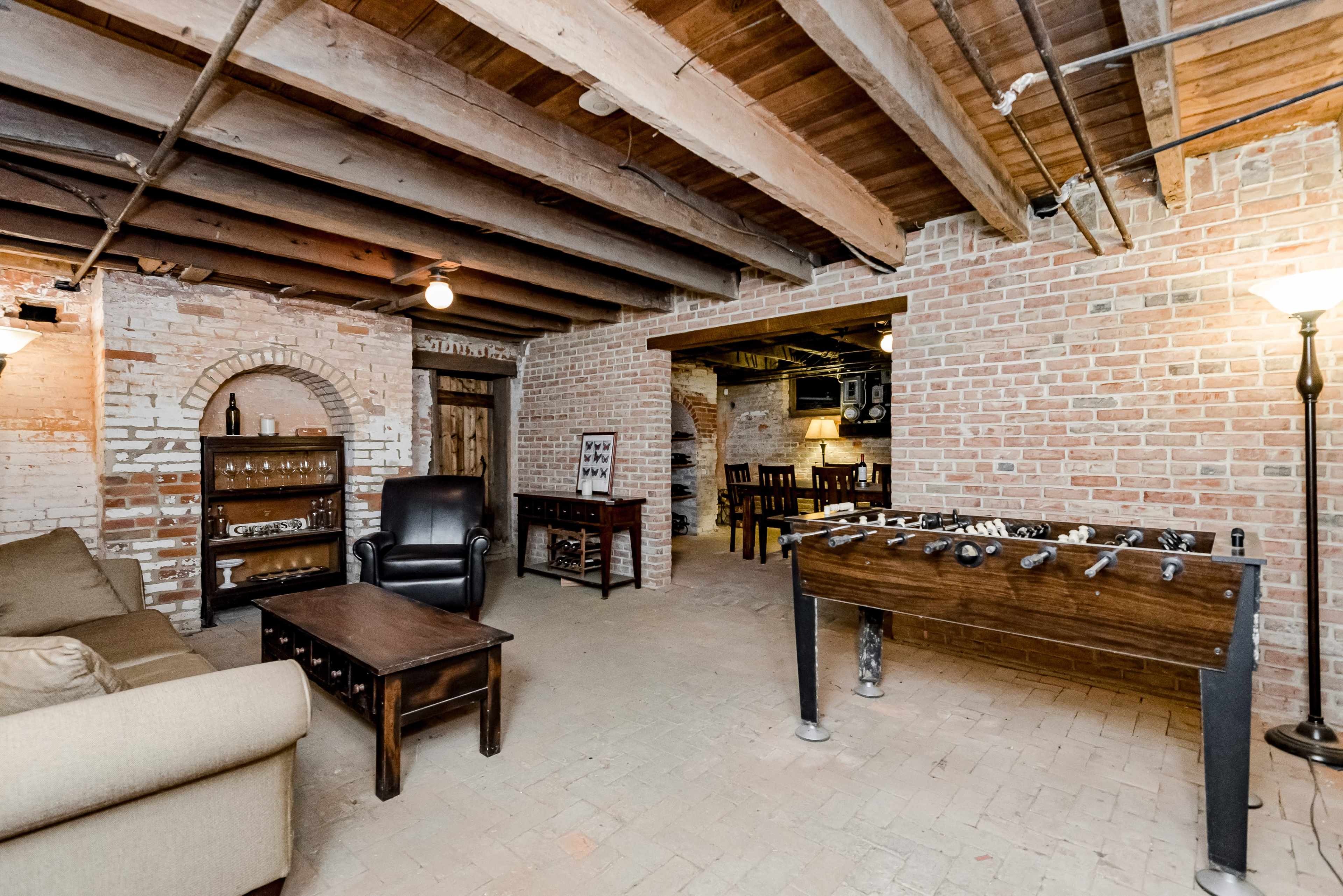The image shows a cozy basement lounge area with exposed brick walls, wooden beams, a sofa, a coffee table, a foosball table, and a small dining area in the background.