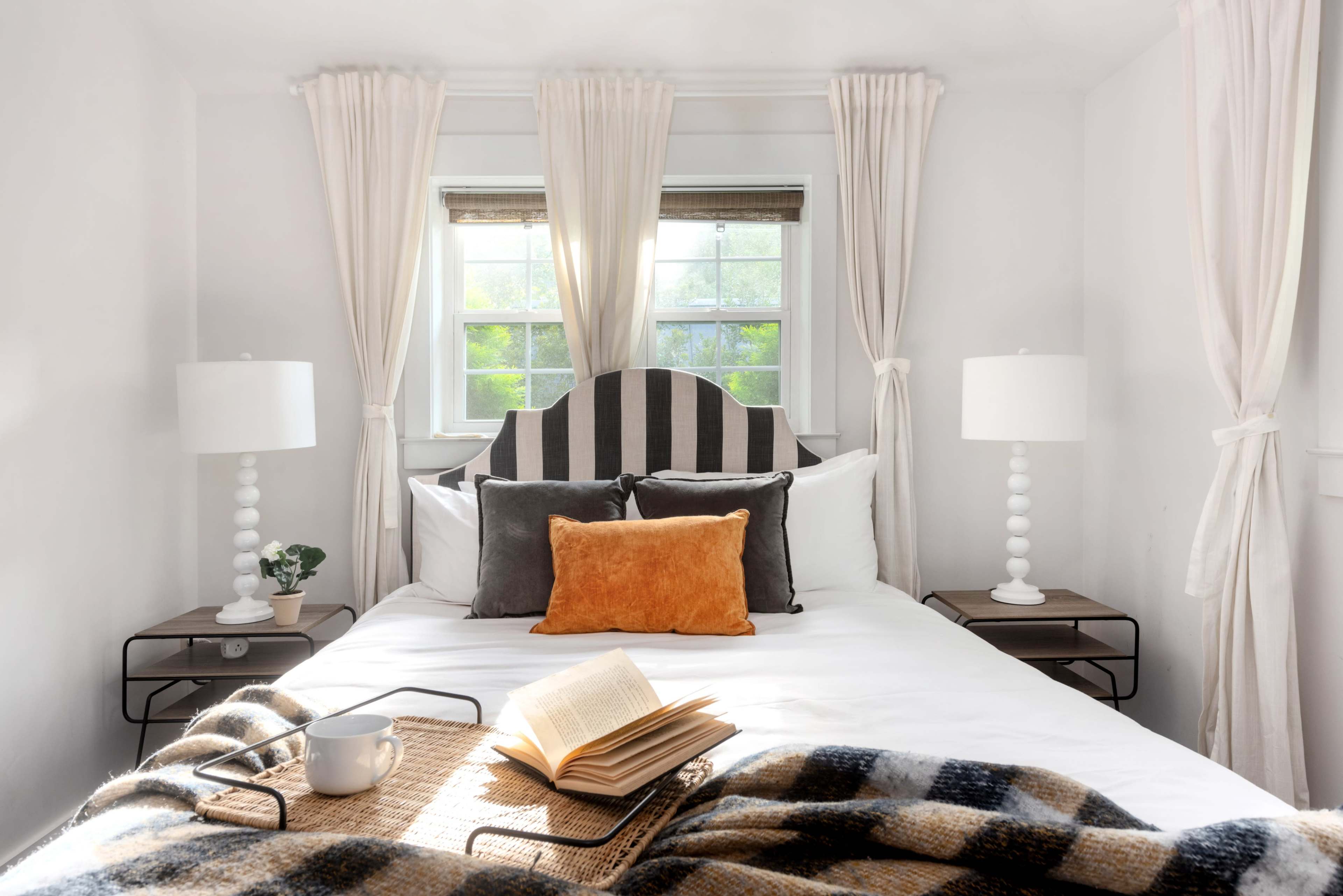 The image shows a cozy bedroom with a bed featuring a striped headboard, flanked by two table lamps and adorned with a blanket and open book on a tray.