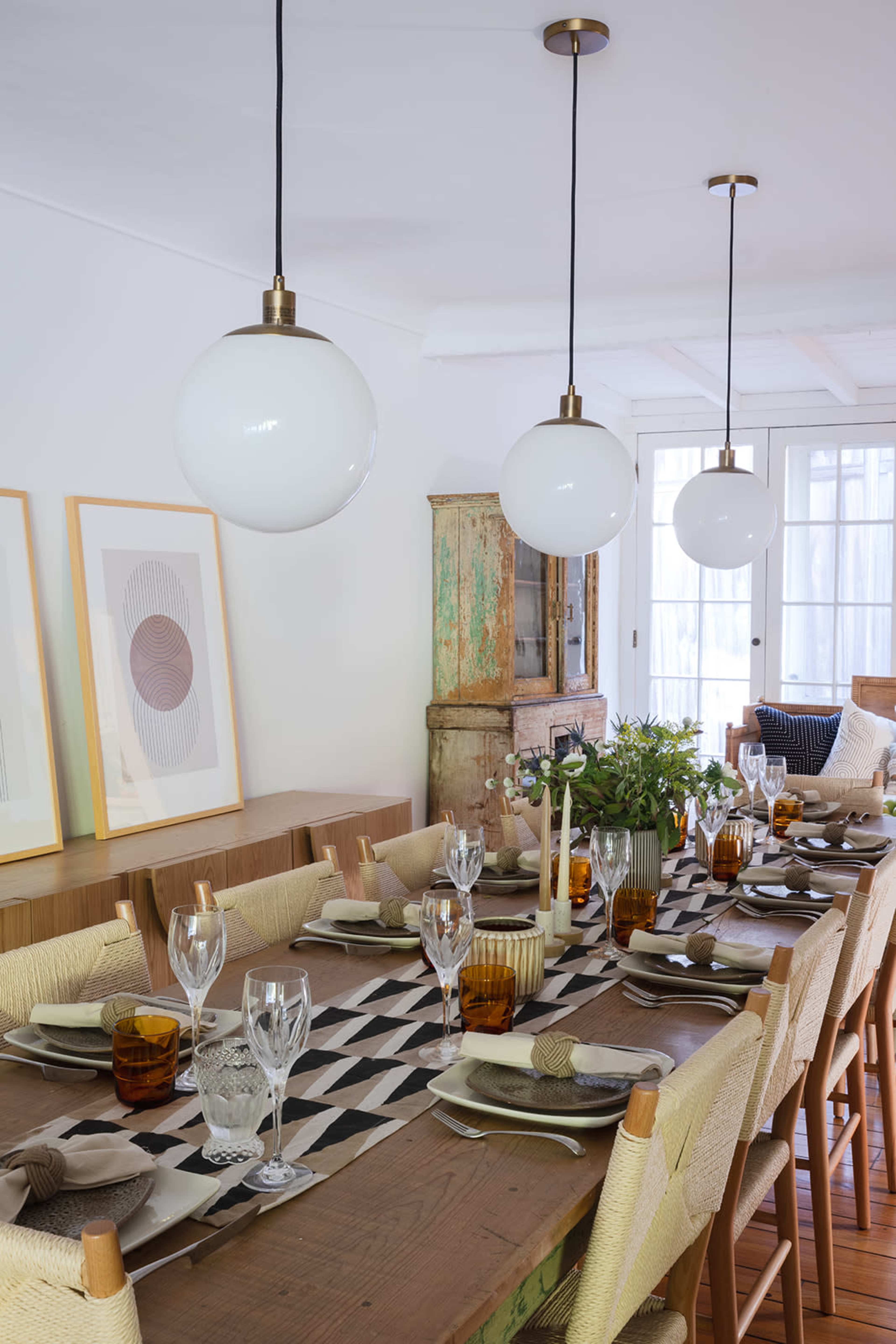 A spacious dining room features a long wooden table set for a meal, surrounded by chairs, with three pendant lights above and large windows in the background.