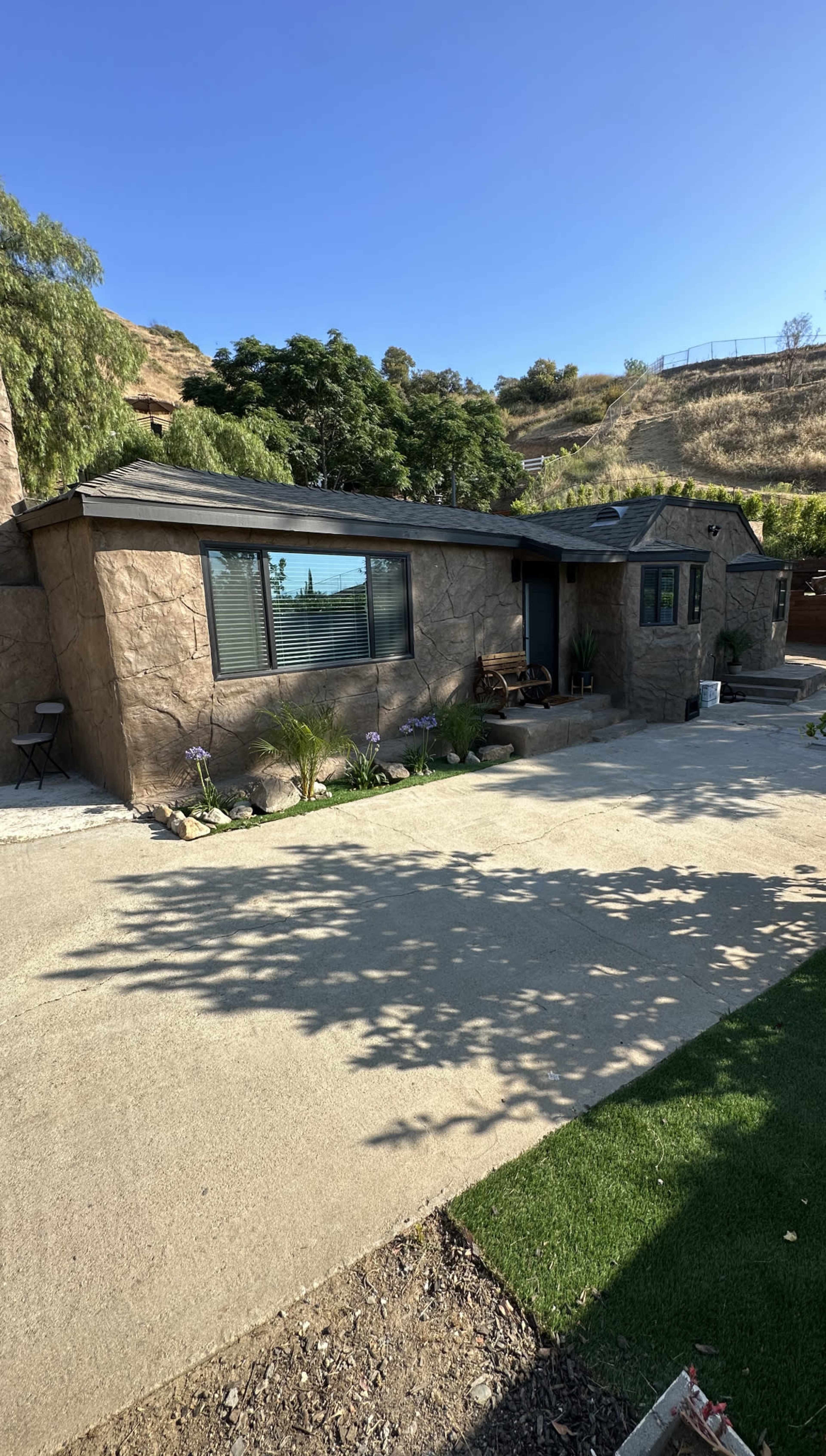 A single-story, stone-textured house with large windows and a porch, set against a hillside with greenery.