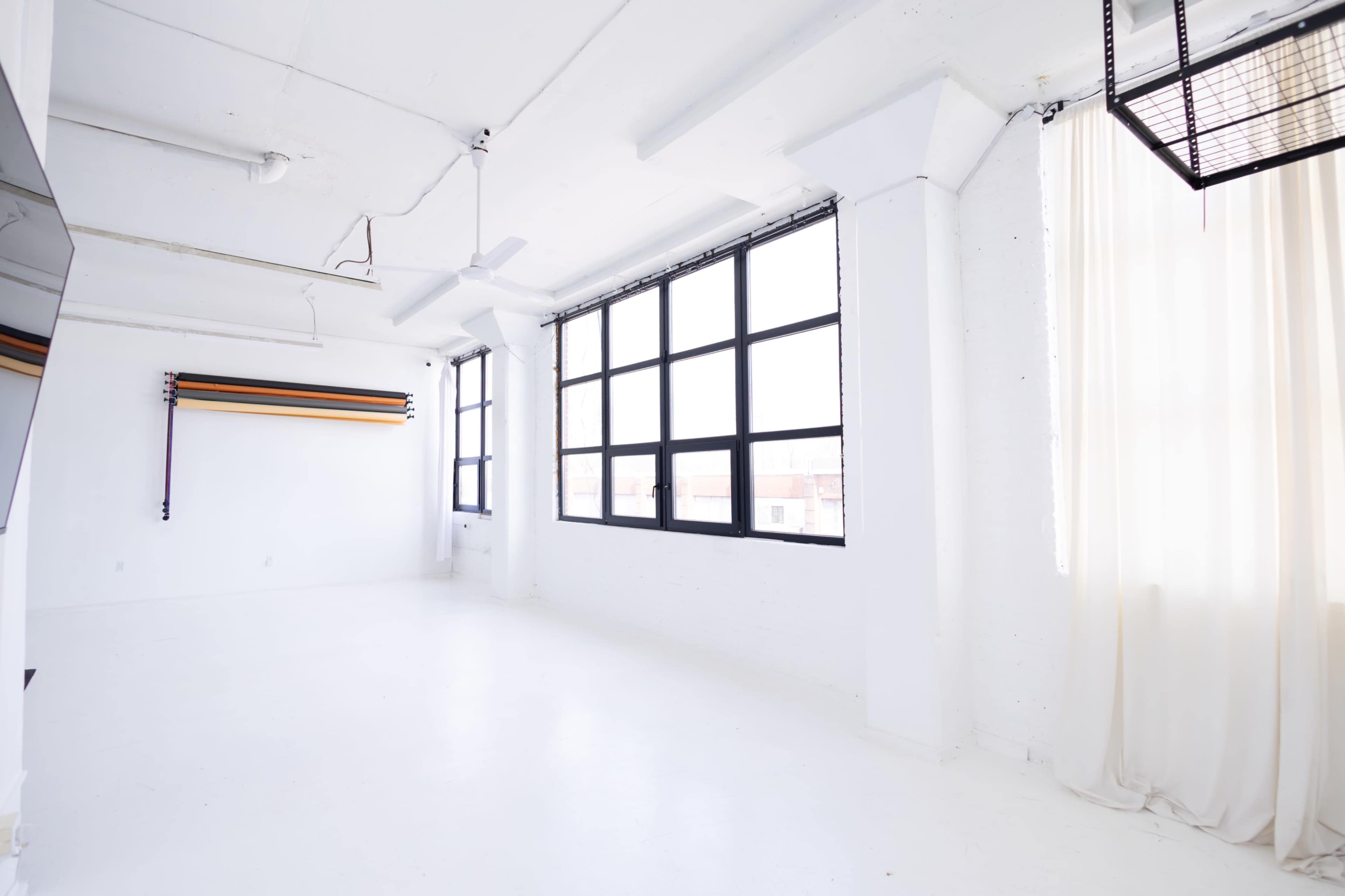 The image shows a bright, empty room with white walls, large windows, and a light-colored floor.