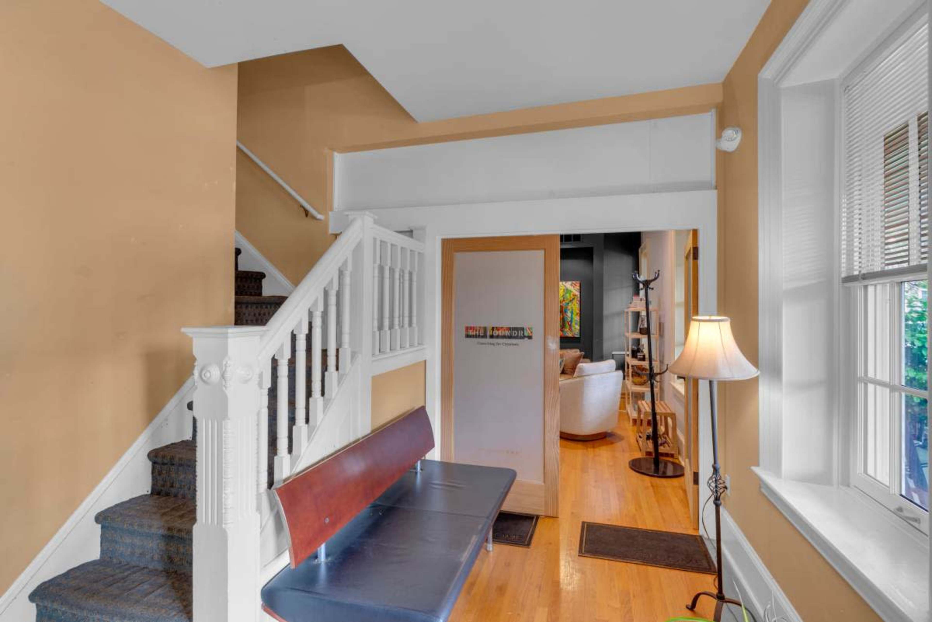 The image shows a residential entryway with a staircase on the left, a bench against the wall, and an opening leading to a living area in the background.