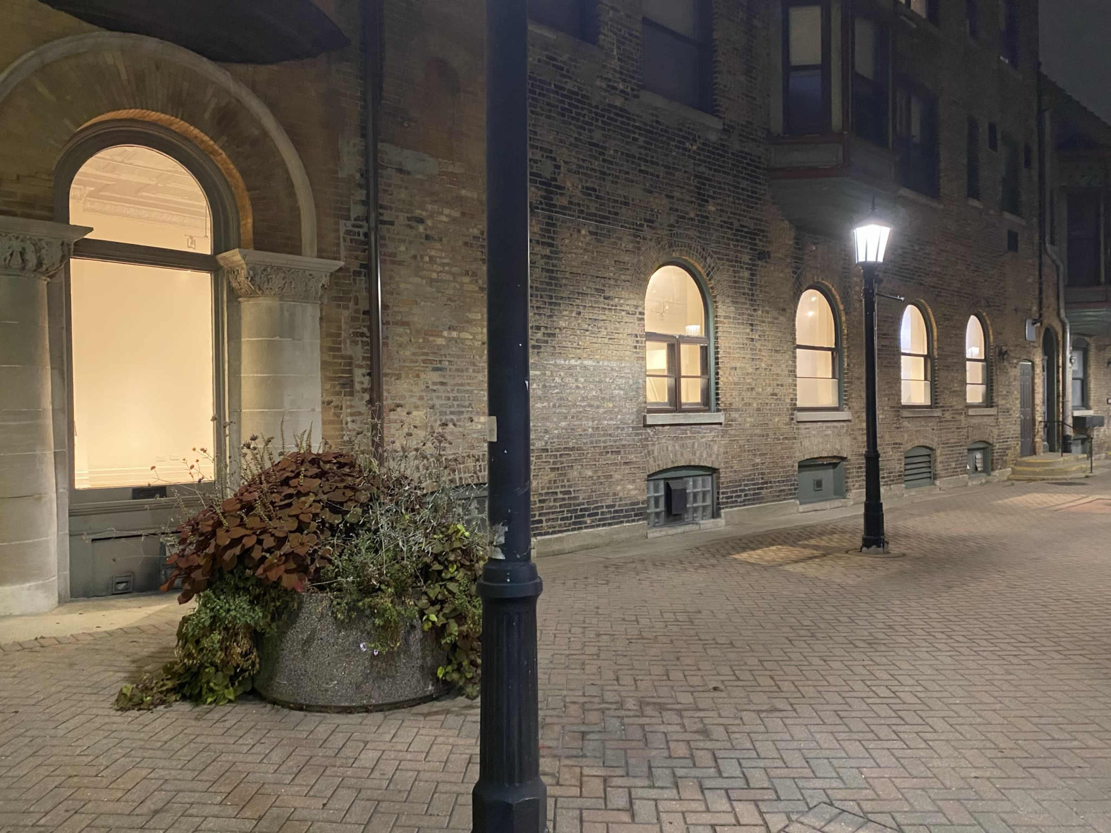 A brick pathway leads alongside a building with arched windows, illuminated by a streetlamp at night.