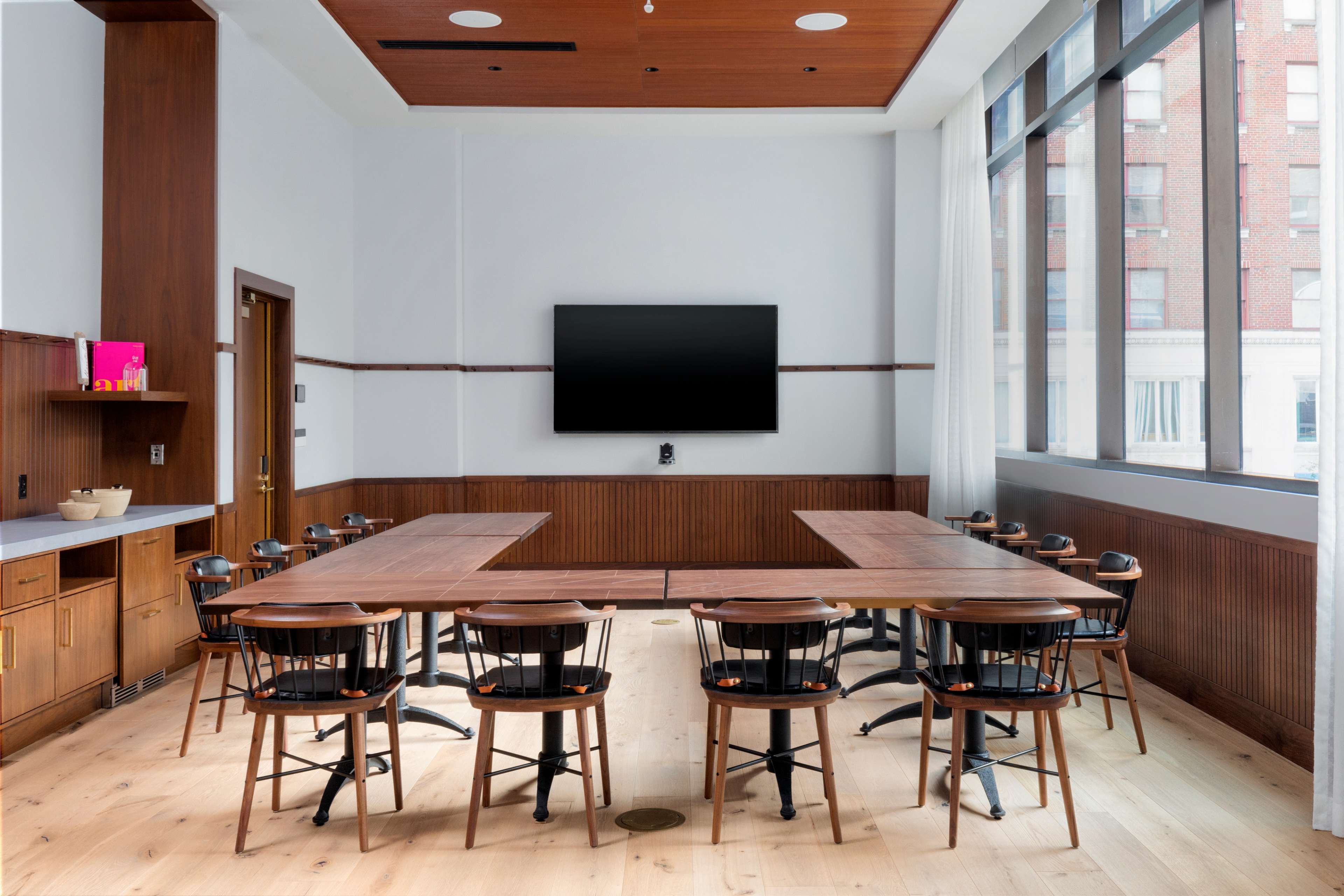 A modern conference room features a lengthy wooden table surrounded by black chairs, with a large screen mounted on the wall and windows allowing natural light.
