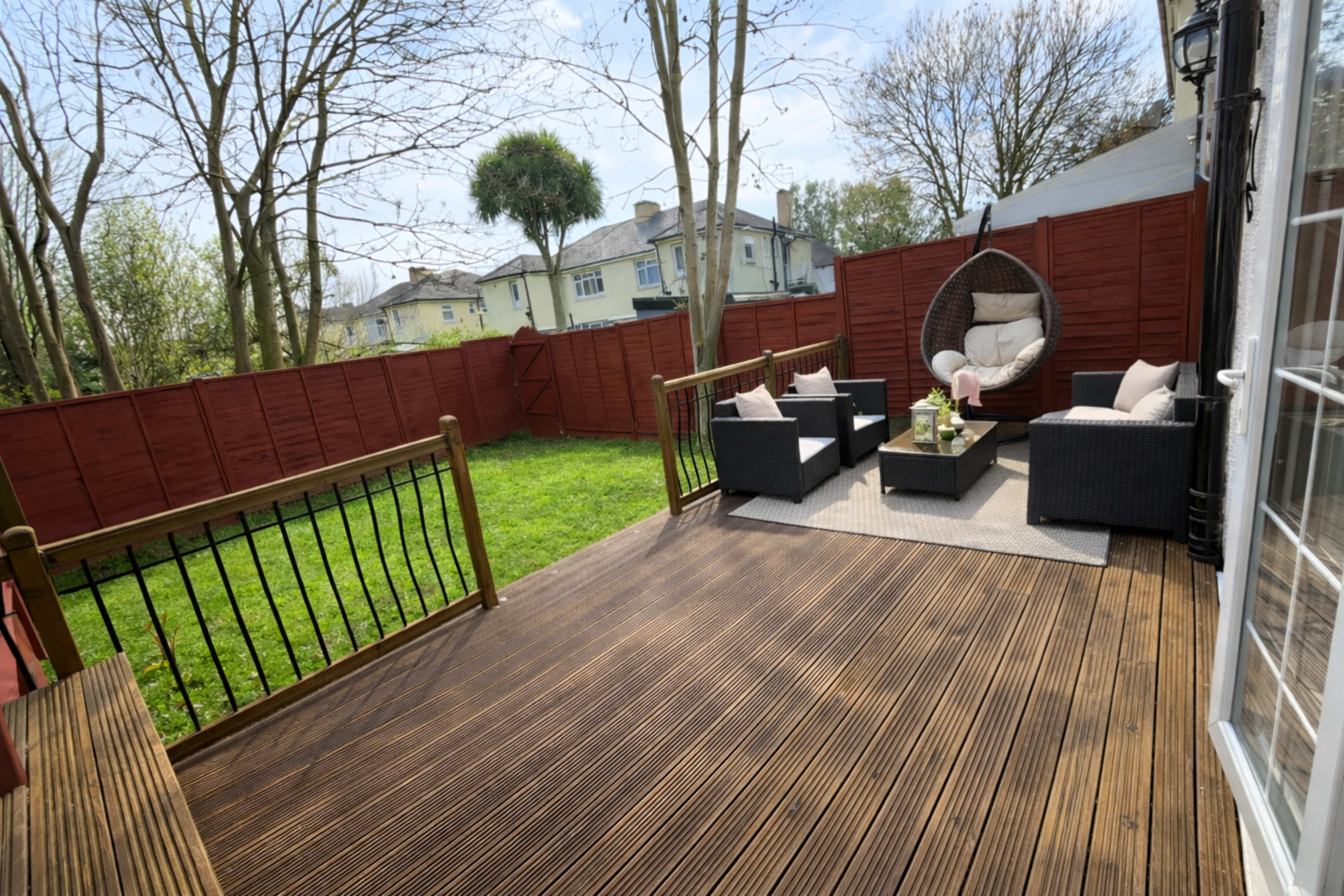 The image shows a wooden deck area furnished with two black wicker chairs, a hanging chair, and a small table, surrounded by a red fence and greenery in the background.