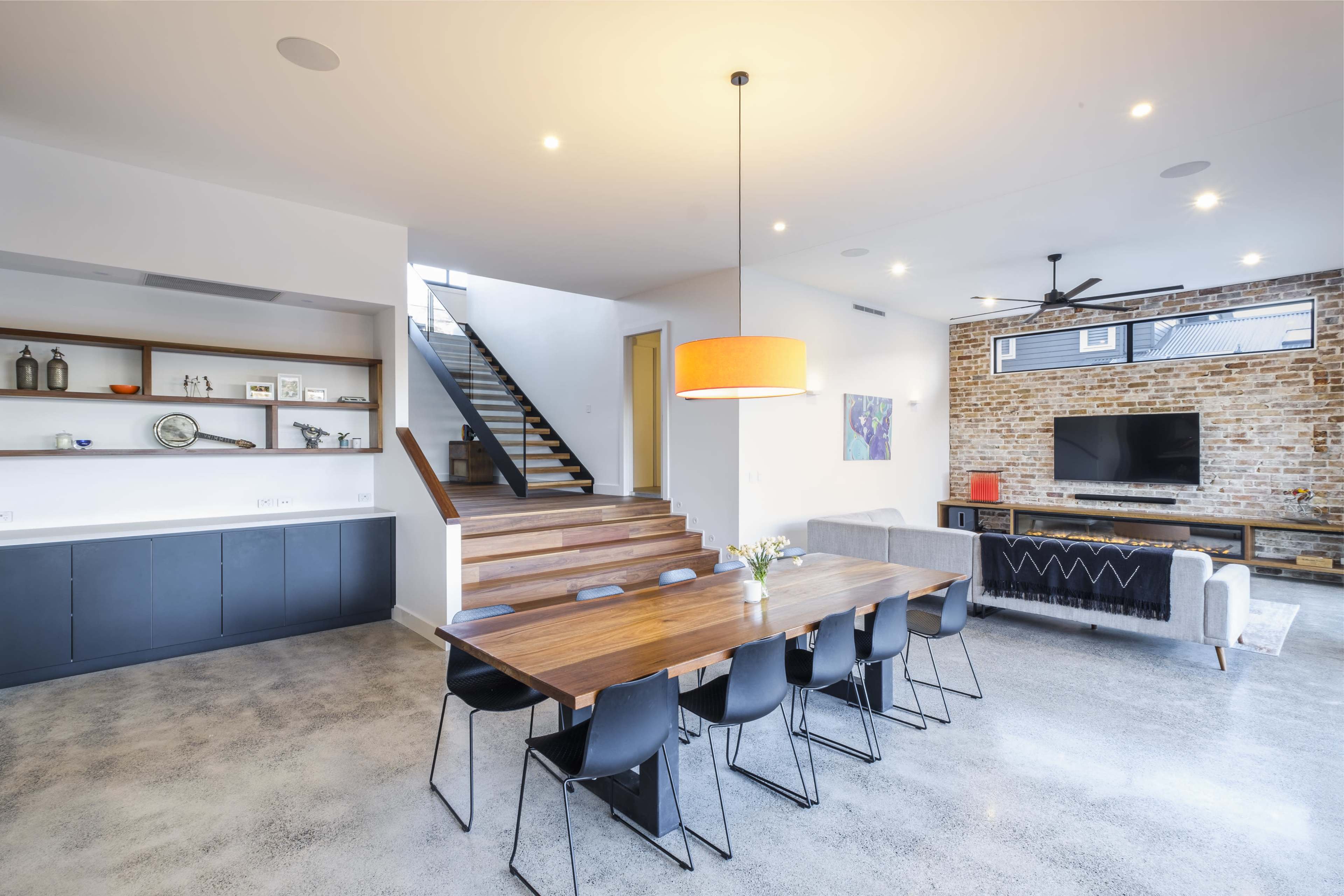 A modern dining area with a wooden table, black chairs, and a staircase leading to an upper level, accompanied by a stone wall and a television setup in the background.