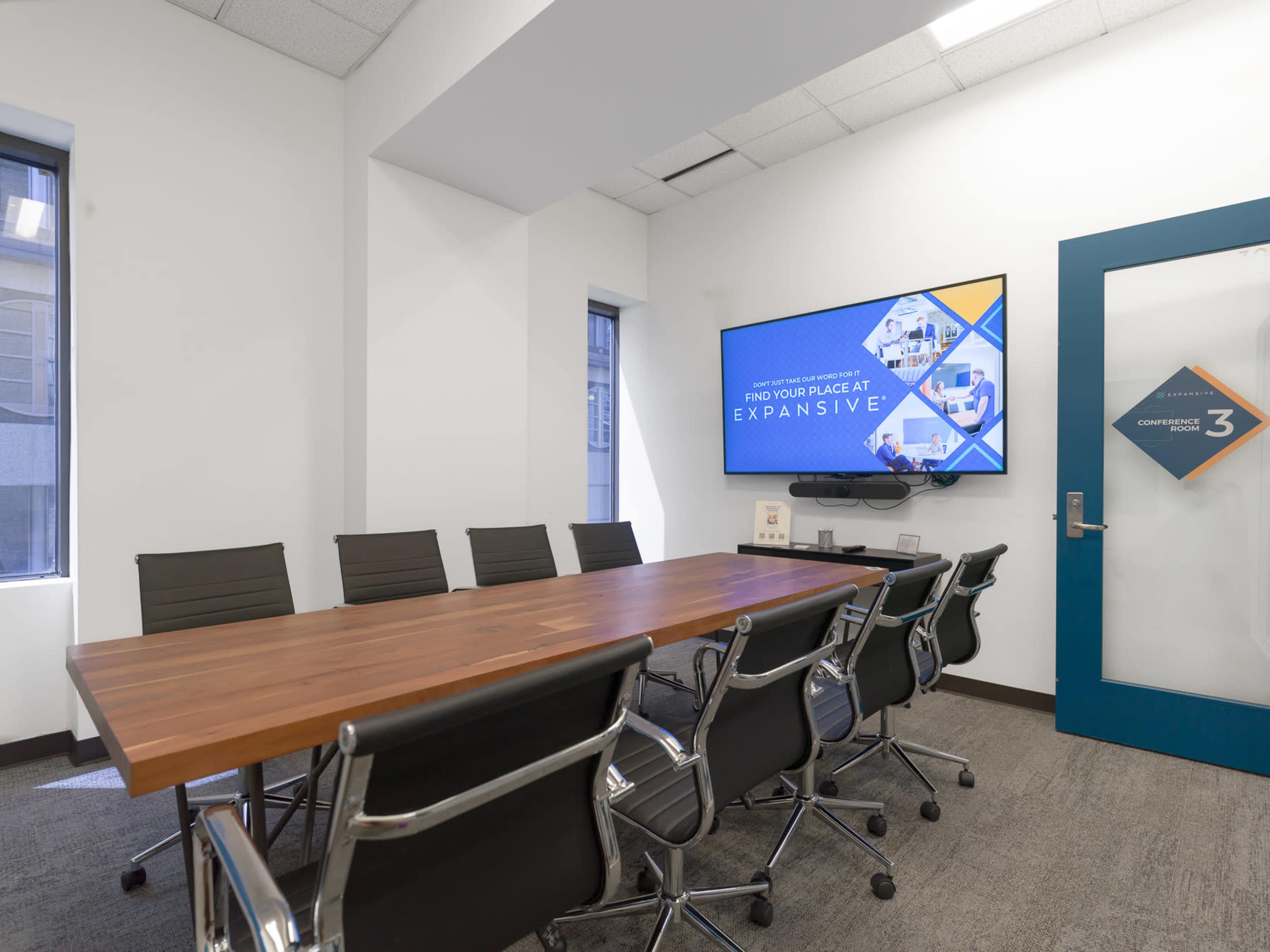 A conference room features a large wooden table surrounded by black chairs, with a screen displaying a presentation on one wall and a glass door labeled "Conference 3."