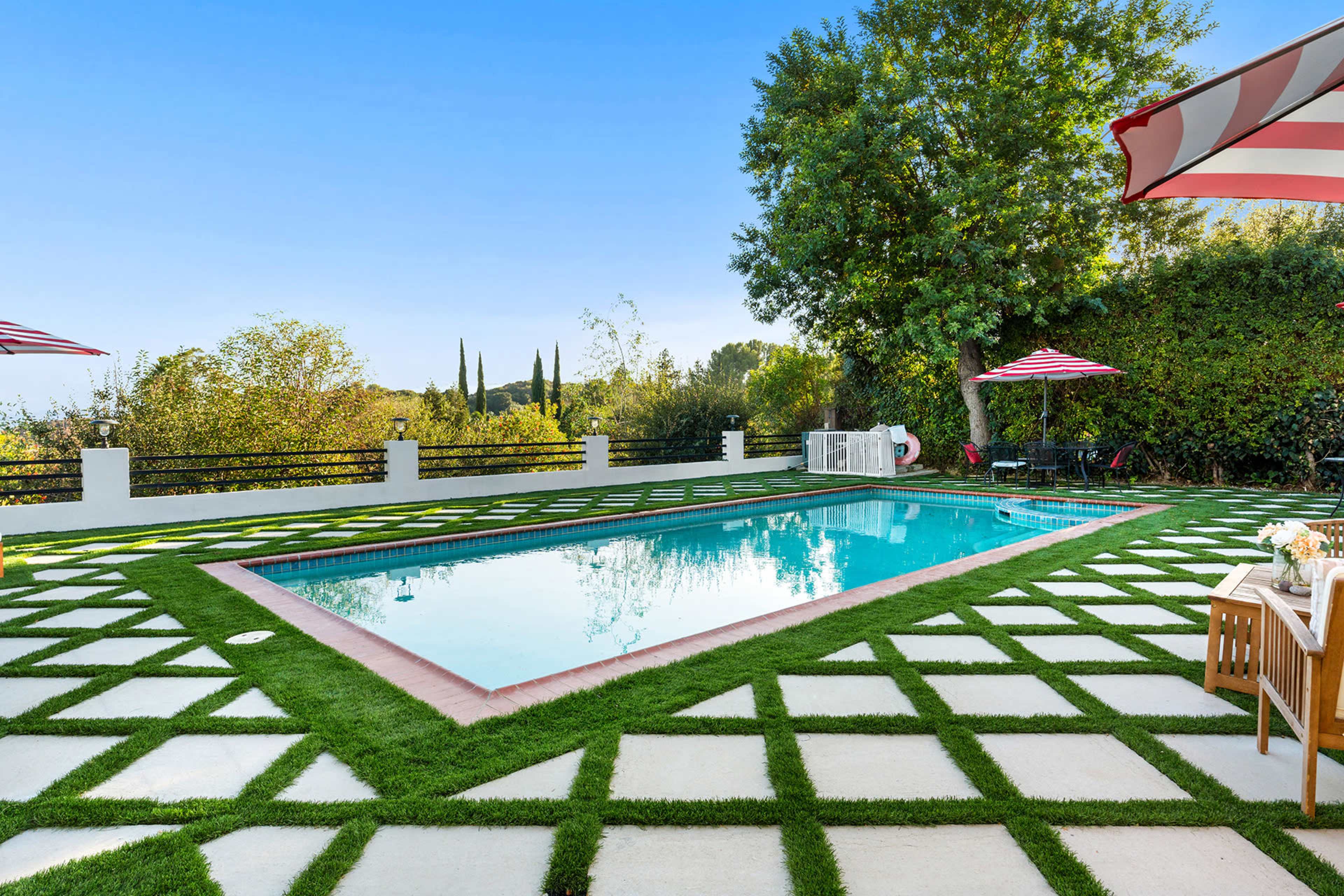 The image features a rectangular swimming pool surrounded by a neatly manicured lawn with geometric stone pavers and decorative umbrellas.