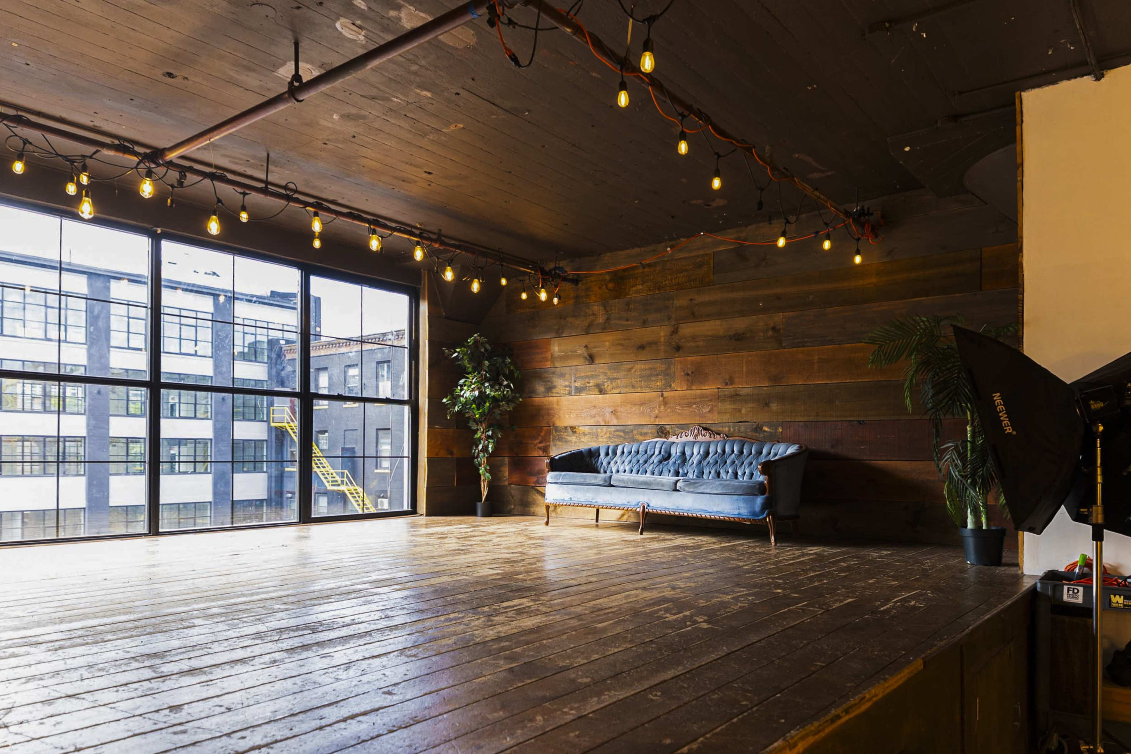 A large, open studio space with wooden floors, a blue sofa, and string lights hanging from the ceiling, illuminated by natural light from large windows.