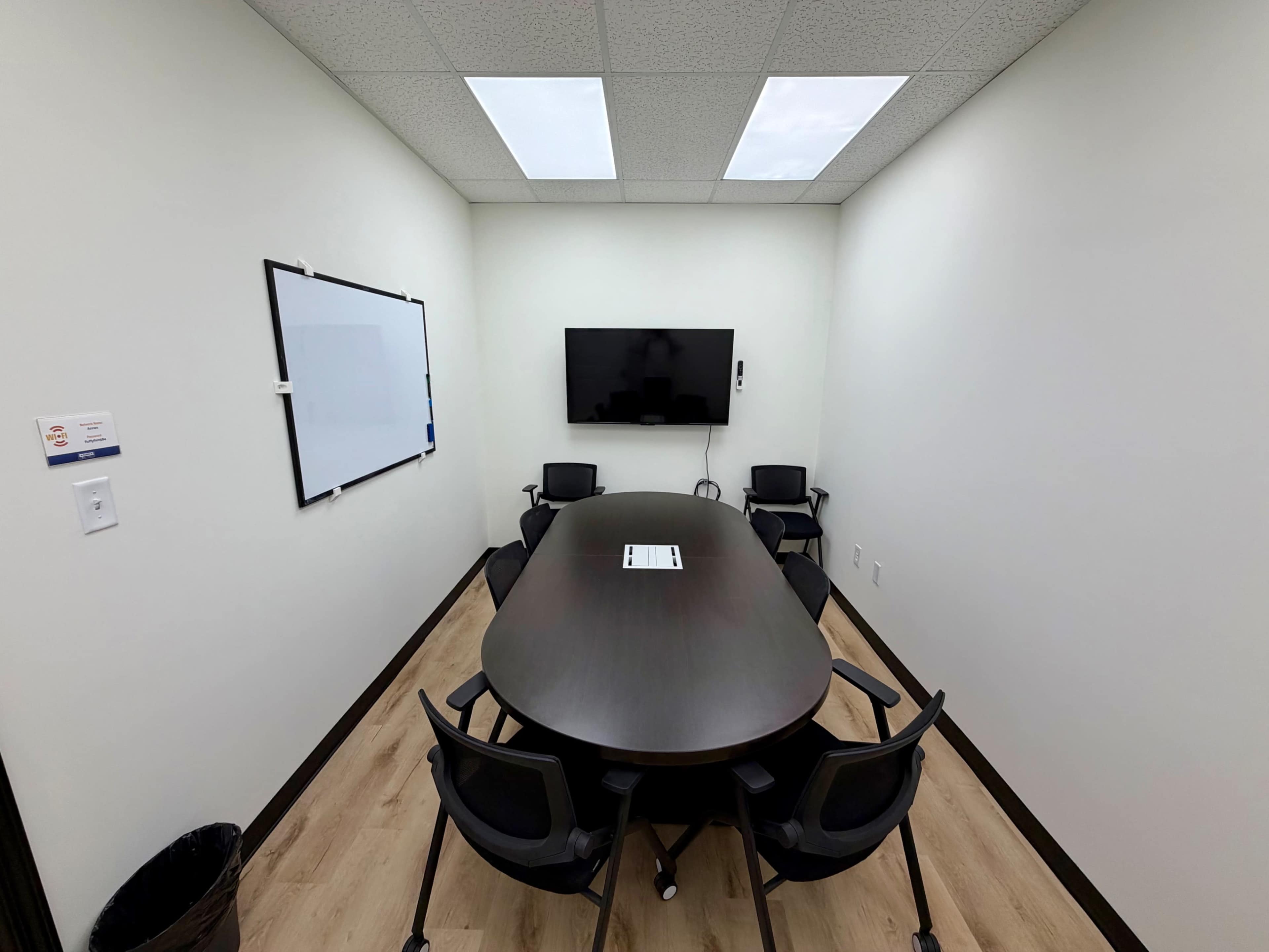 The image shows a small conference room with a round table, six black chairs, a wall-mounted television, and a whiteboard.