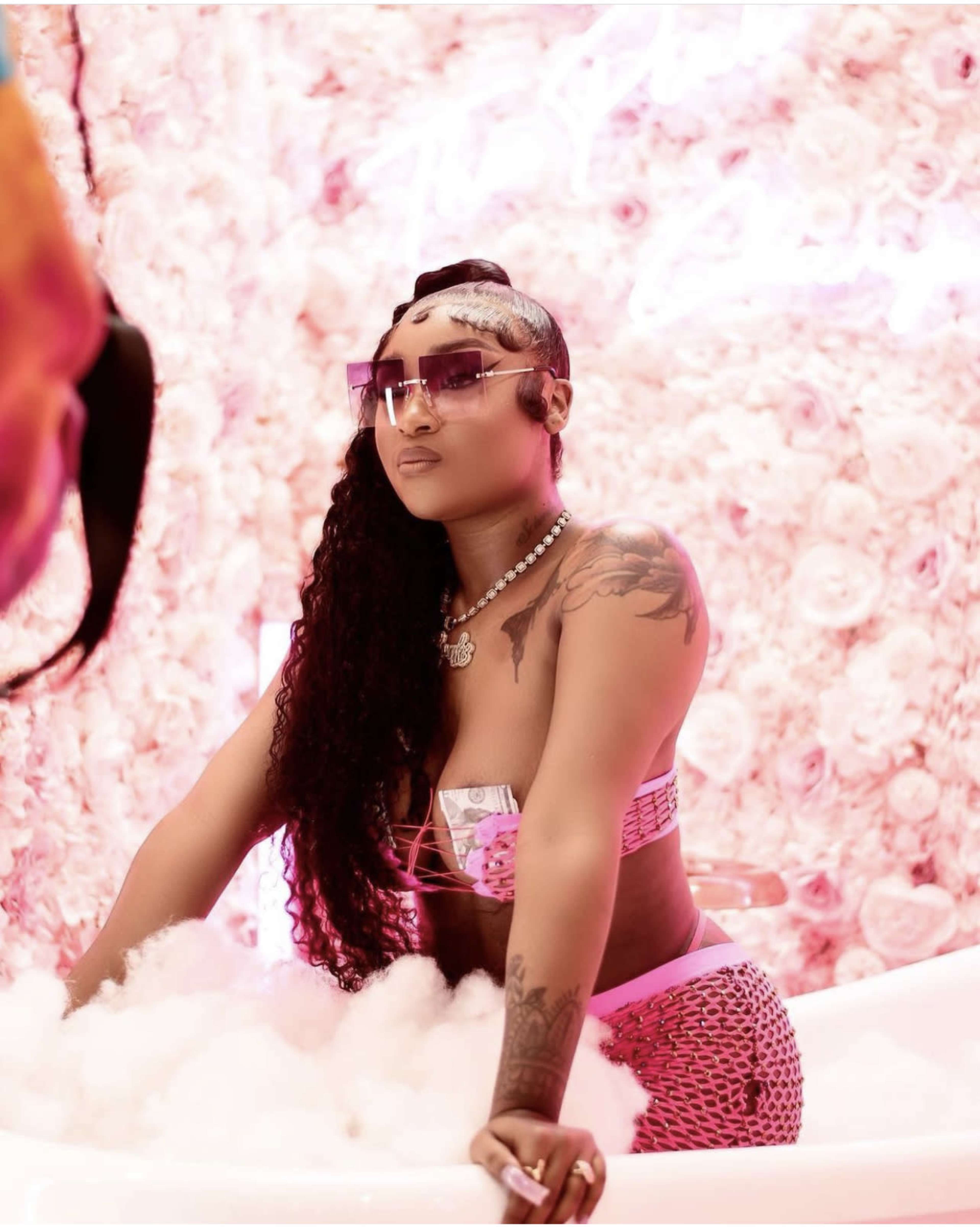 A person with long hair and sunglasses poses in a bathtub surrounded by pink flowers and a cloud of bubbles.