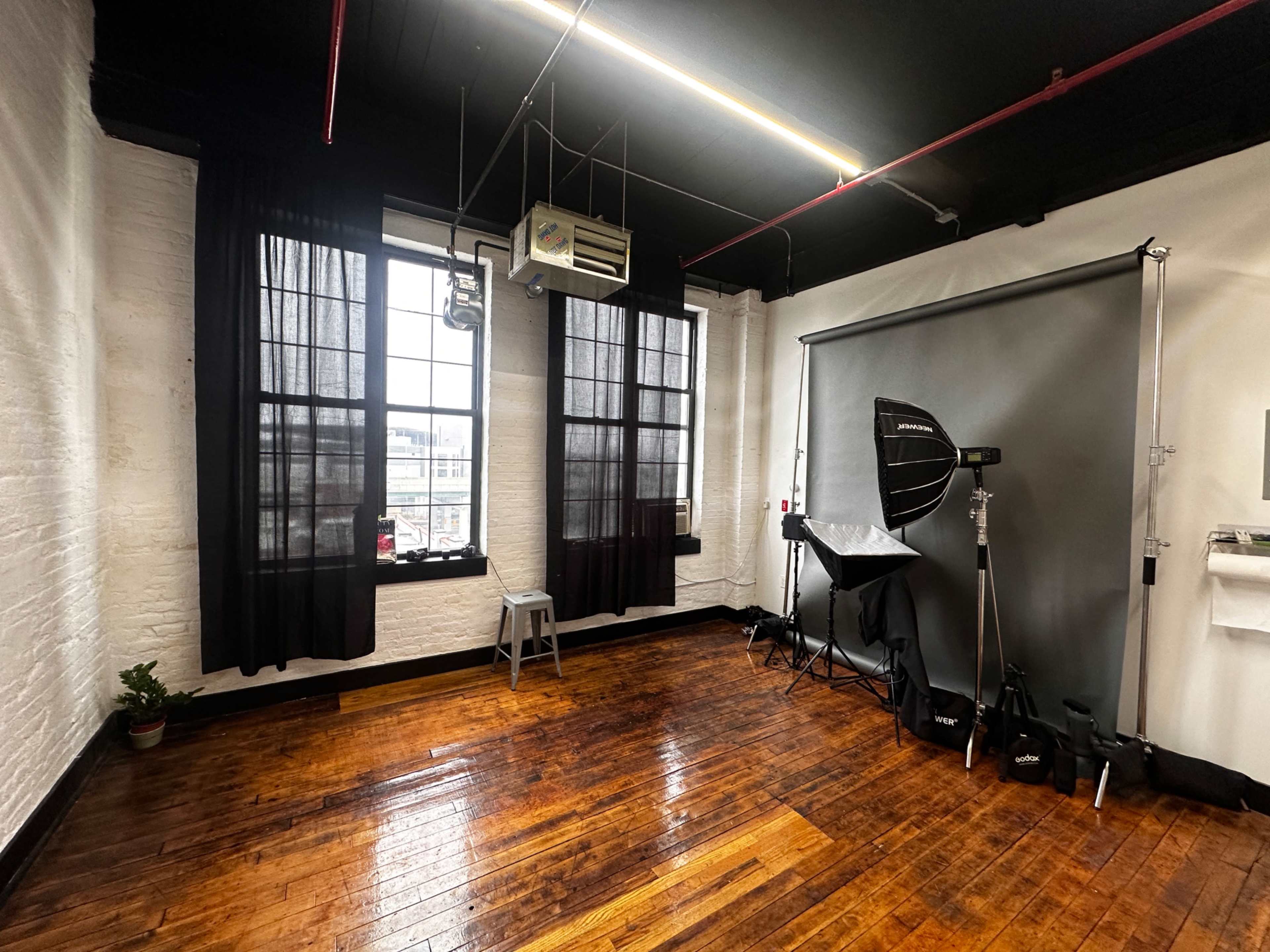 A spacious photography studio features wooden floors, large windows with black curtains, and a backdrop setup with lighting equipment.