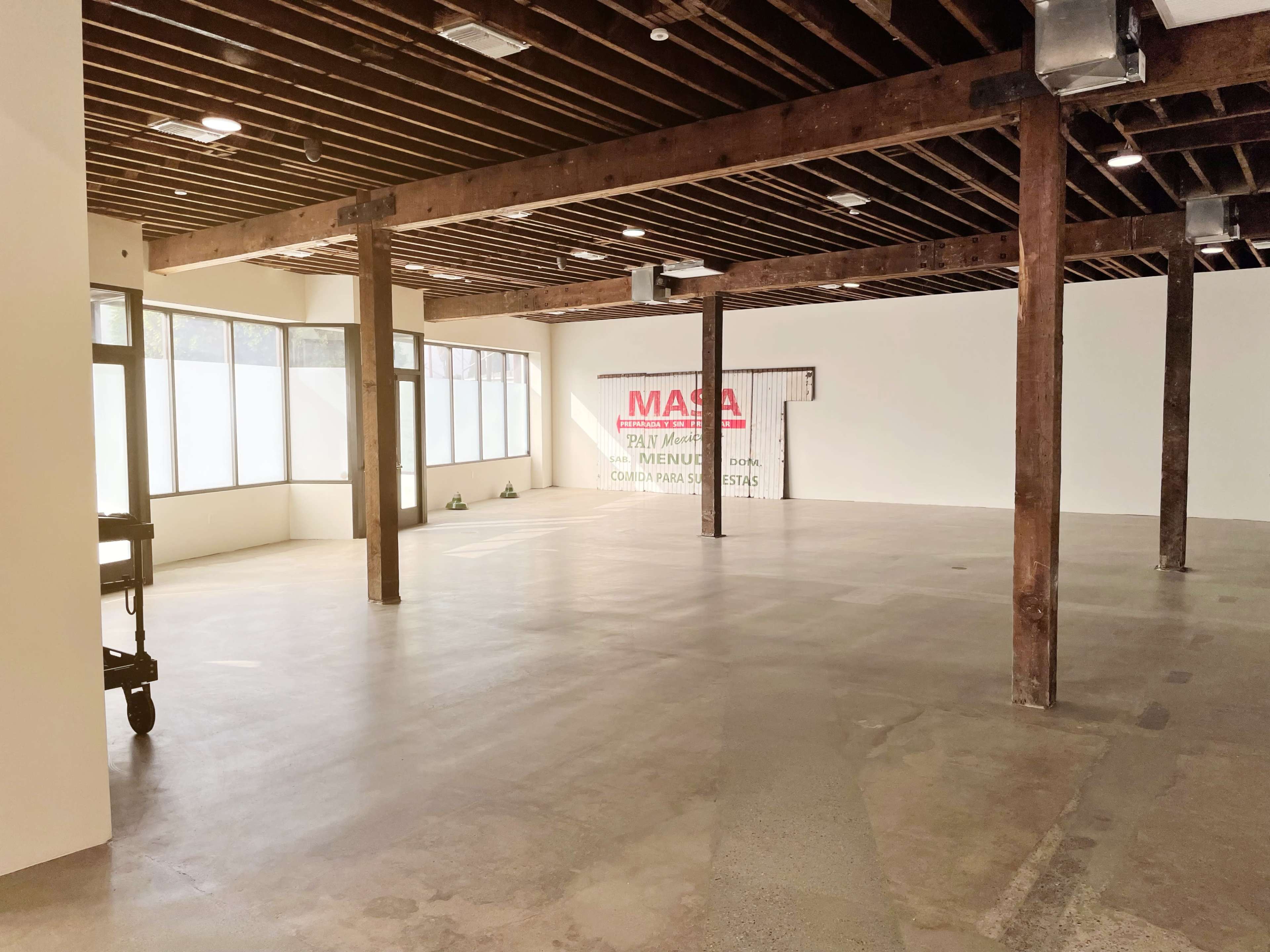 An empty room with exposed wooden beams, large windows, and a concrete floor.