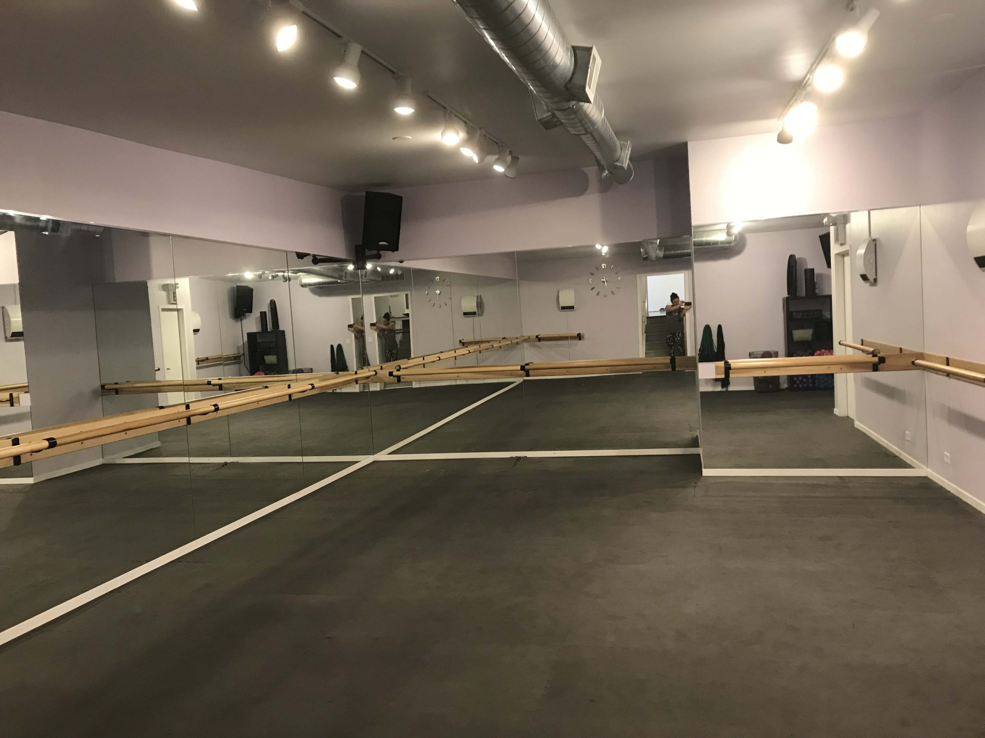 A dance studio with mirrored walls, a ballet barre along one side, and concrete flooring.