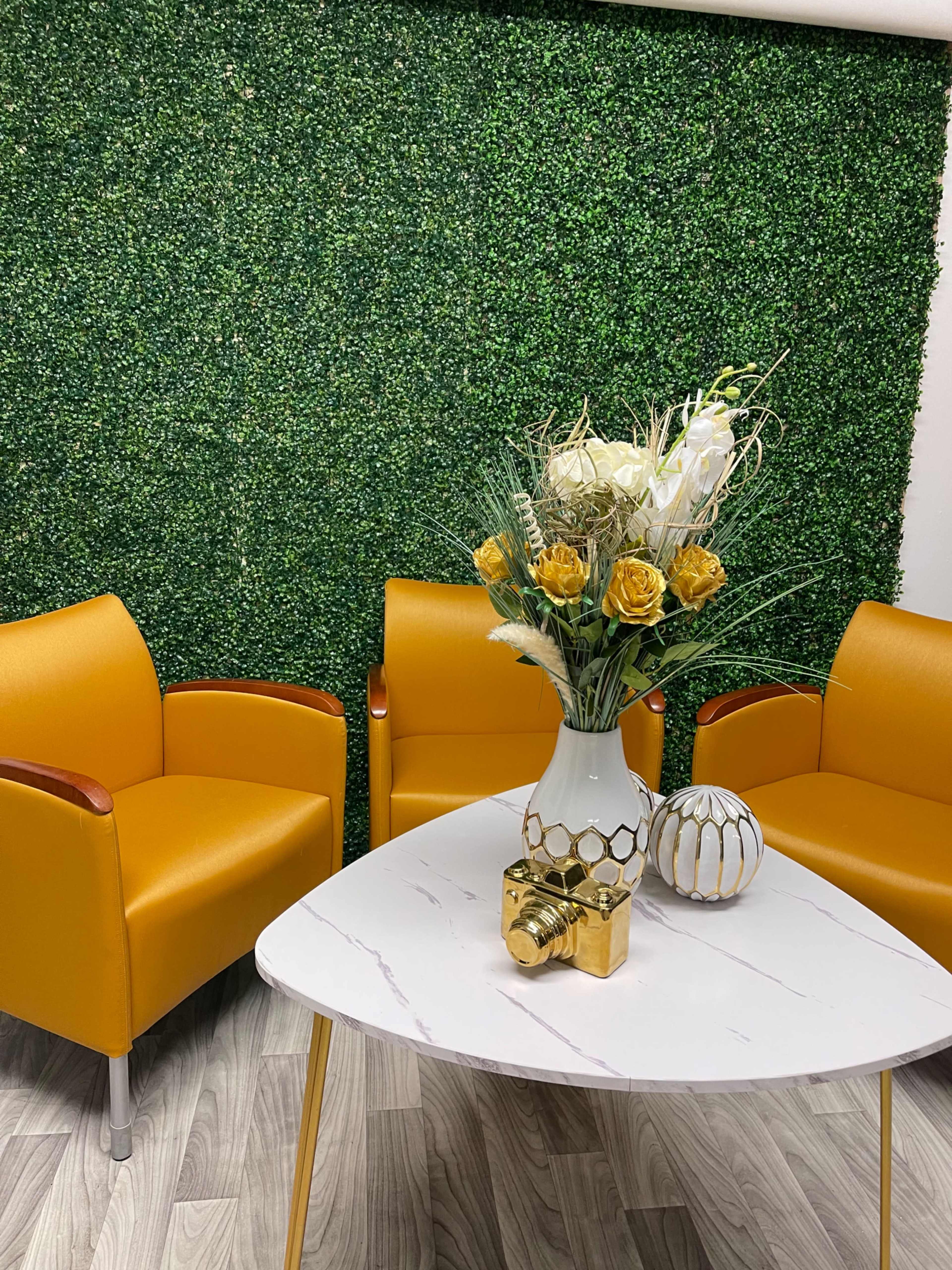 The scene features two yellow chairs and a marble-topped table with a floral arrangement on a green backdrop of artificial foliage.