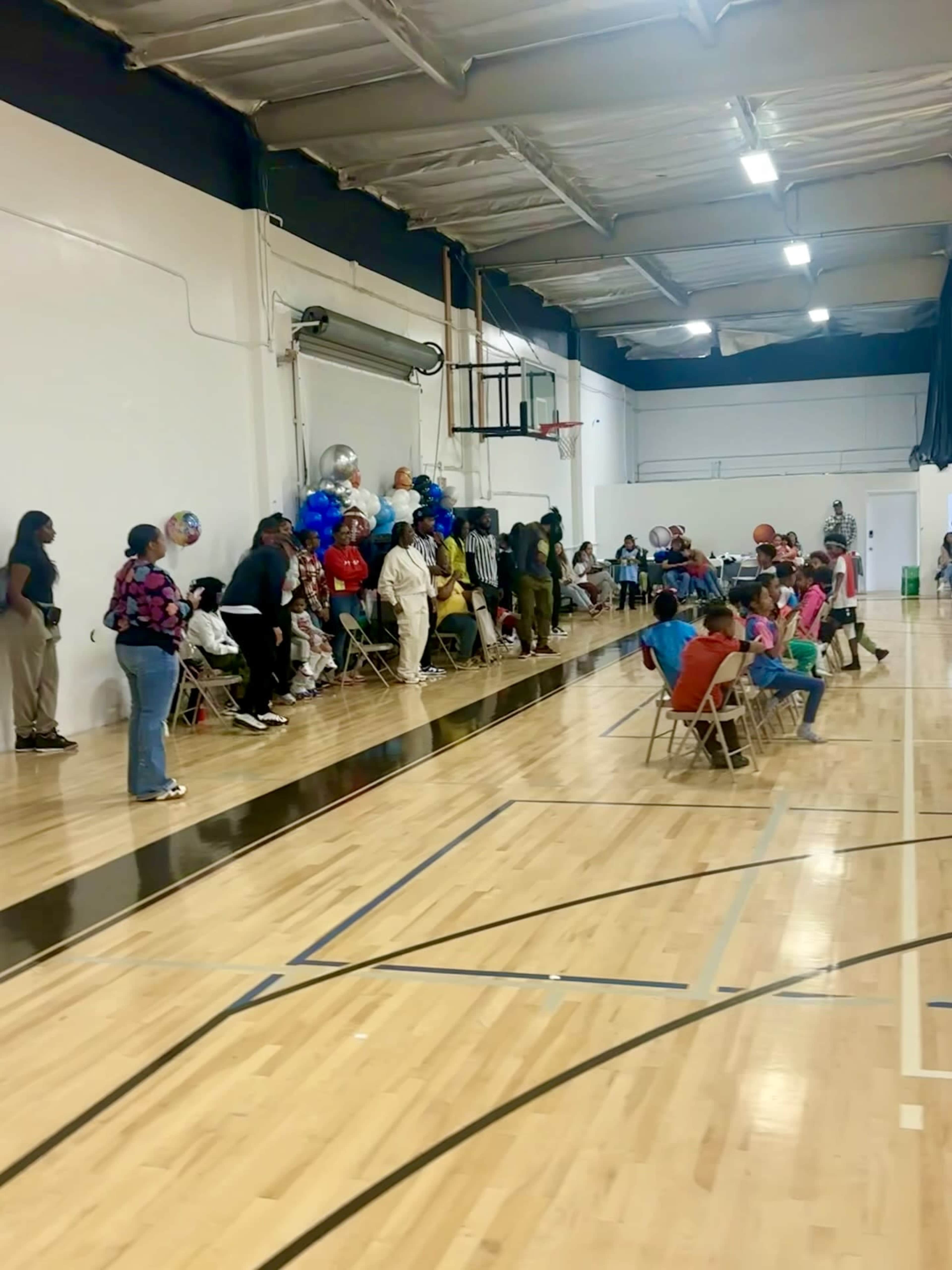 A group of people is seated along the sides of a gymnasium while others are gathered at tables in the center, decorated with balloons and colorful attire.