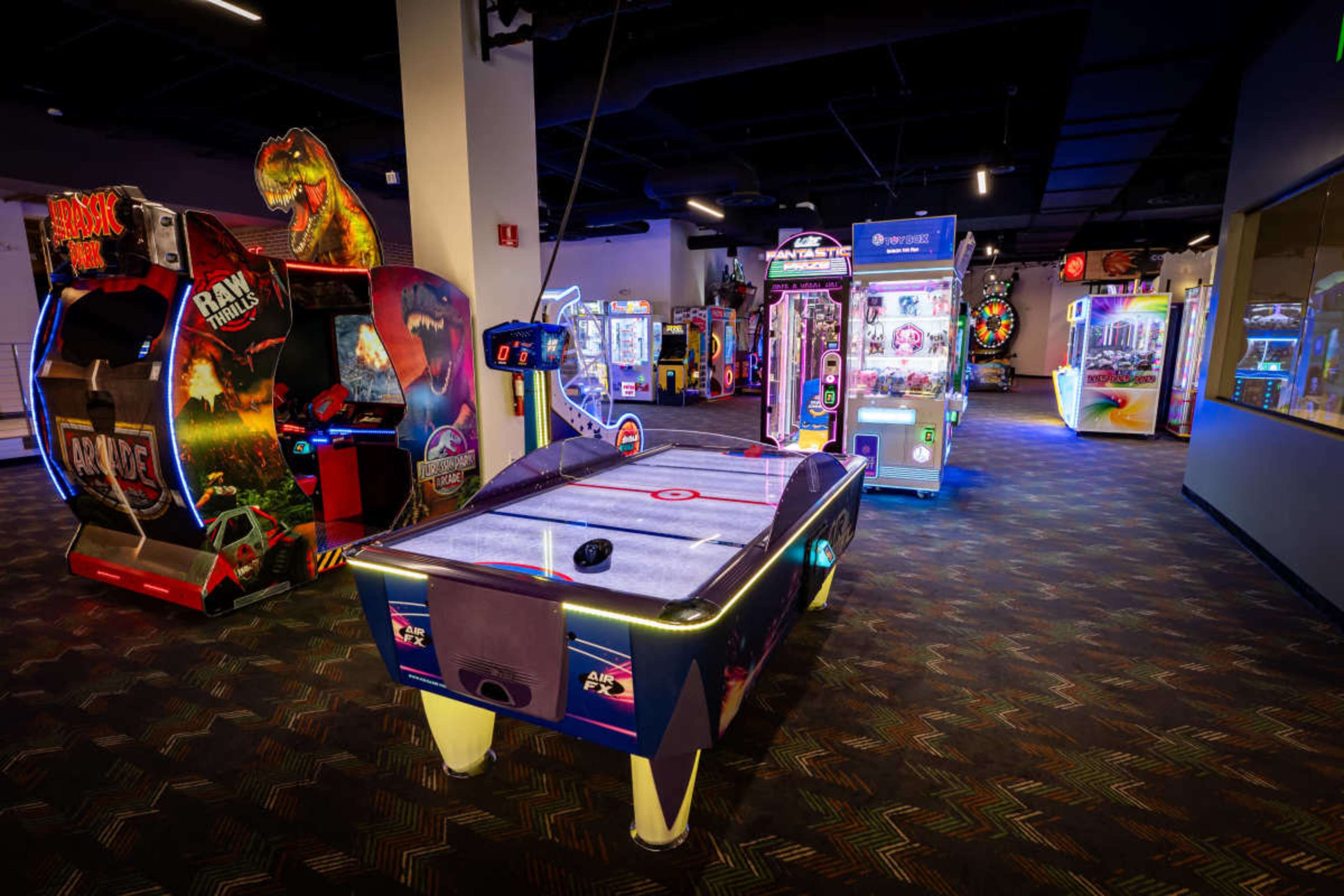 The image shows an arcade featuring various gaming machines, including air hockey, claw machines, and classic arcade games set in a dimly lit space.