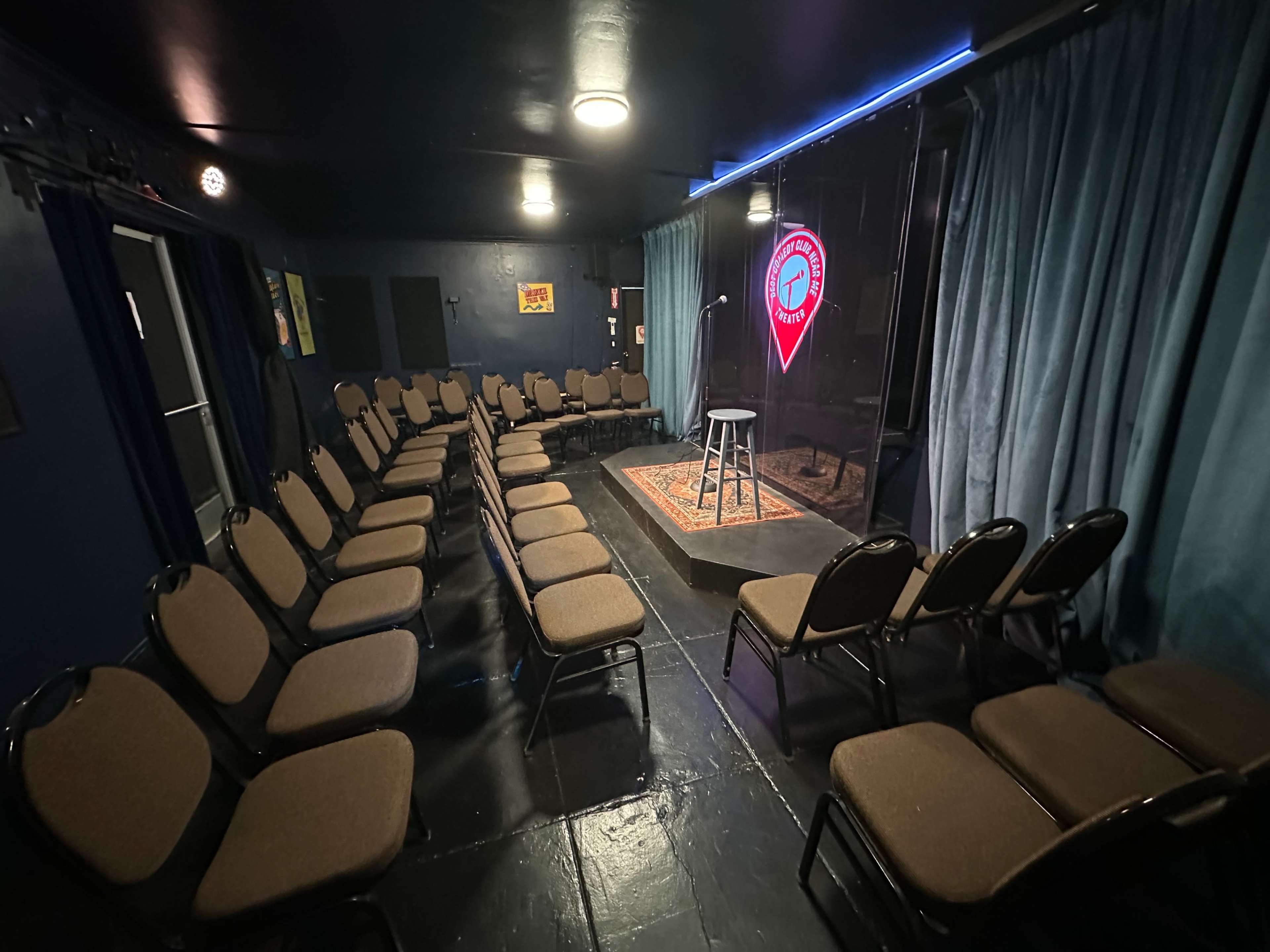 The image shows a small comedy stage with a single stool and a neon sign, facing rows of seated chairs arranged in a semicircle.