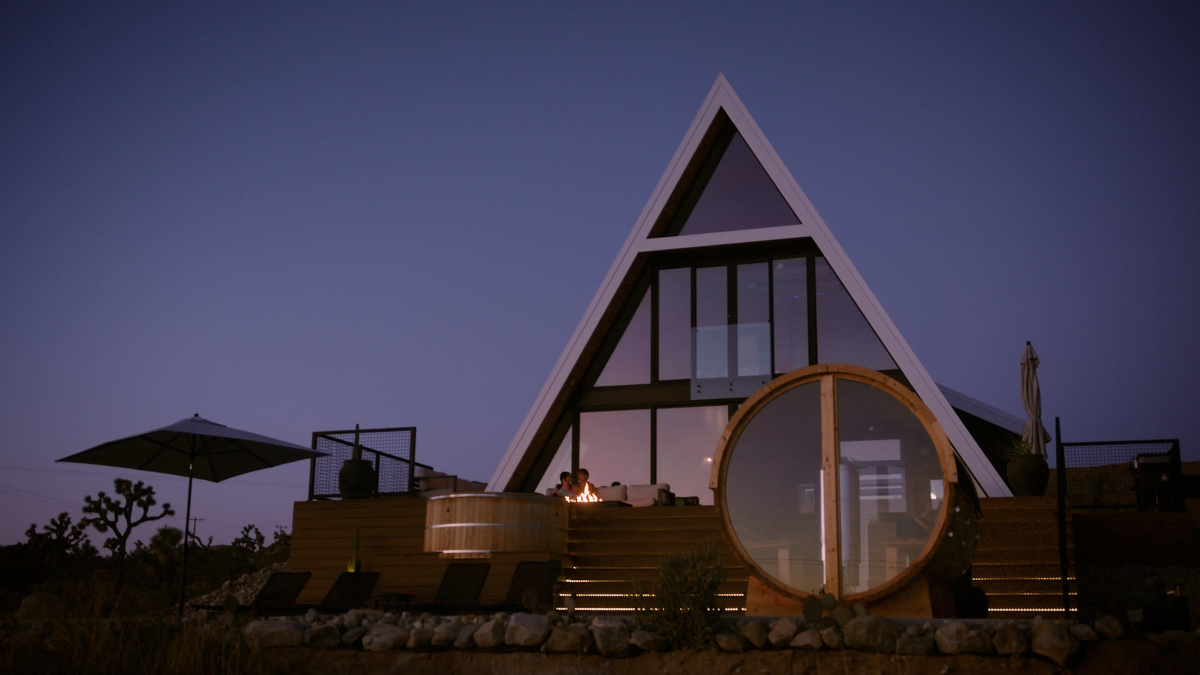 A modern A-frame house with large windows and a circular hot tub is set against a twilight sky.