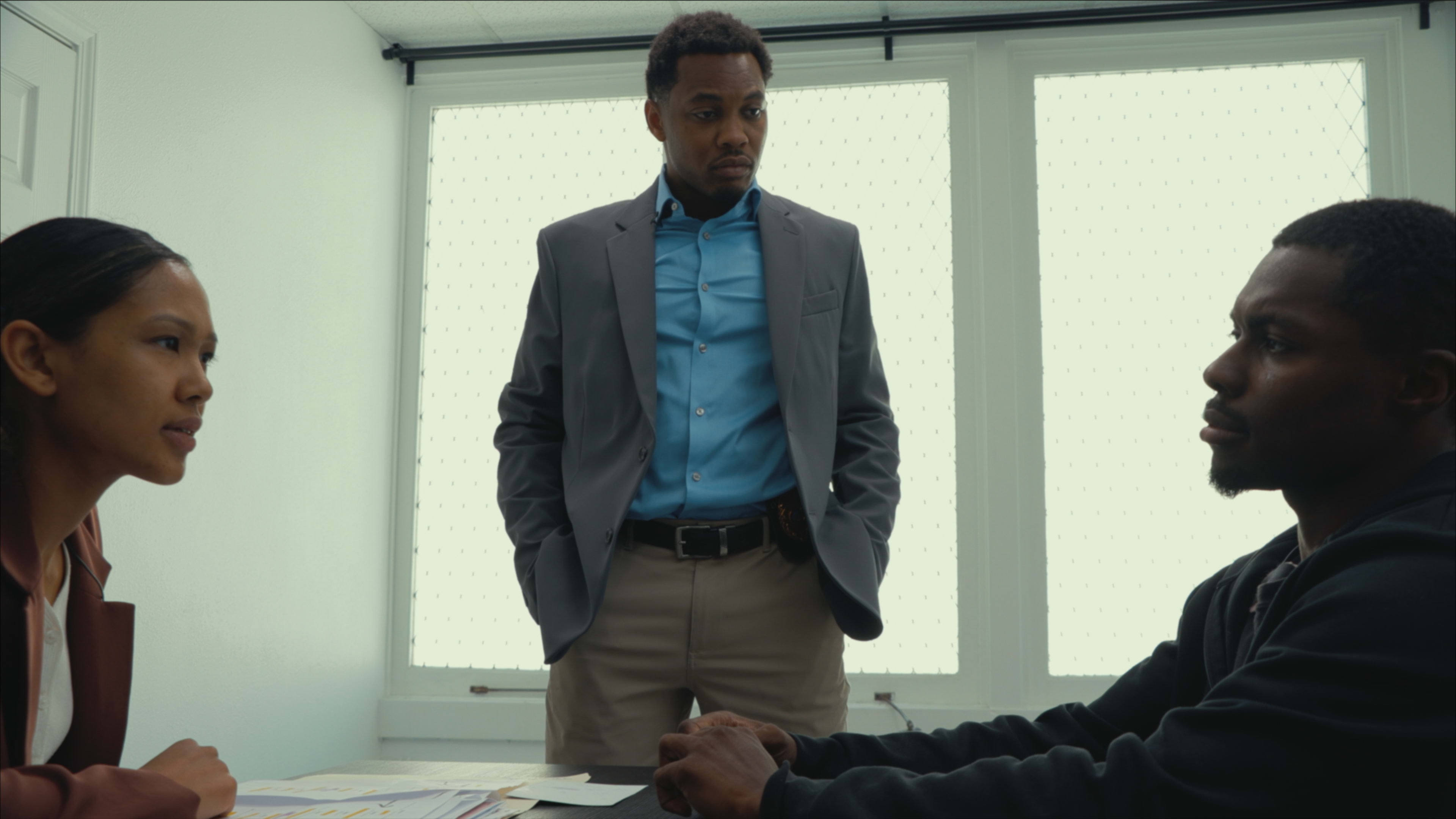 A man in a suit stands with his hands in his pockets while looking down at another man seated at a table, and a woman sits next to the table observing the interaction.