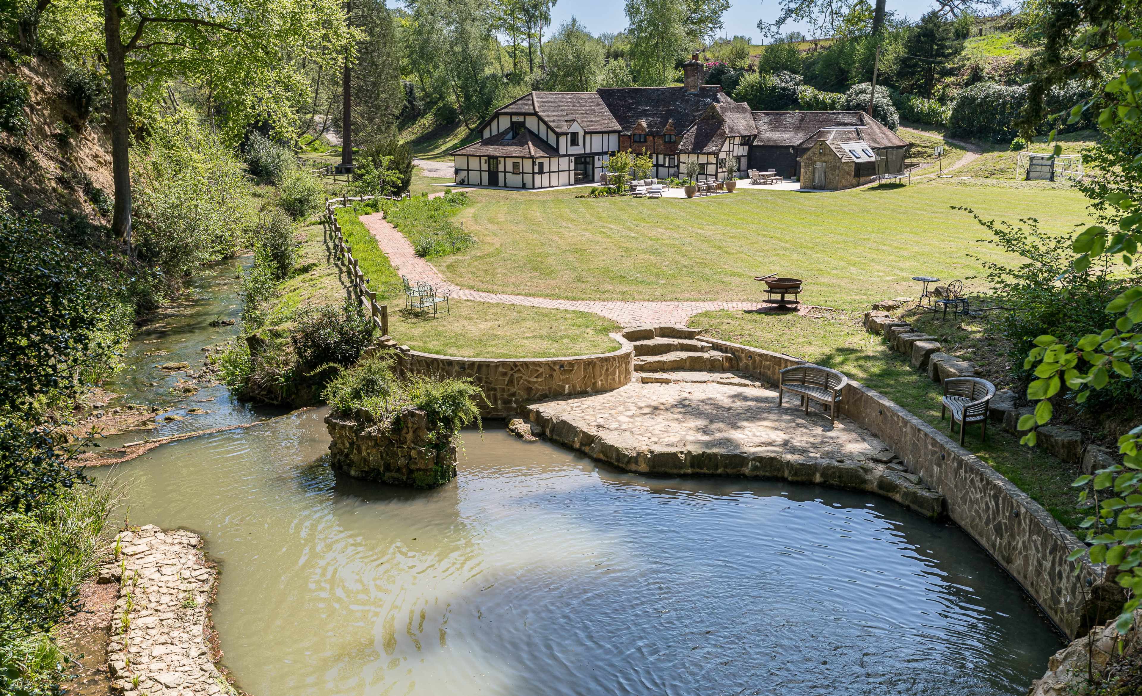 A picturesque landscape features a charming house beside a flowing stream, with manicured lawns and stone pathways leading to seating areas.
