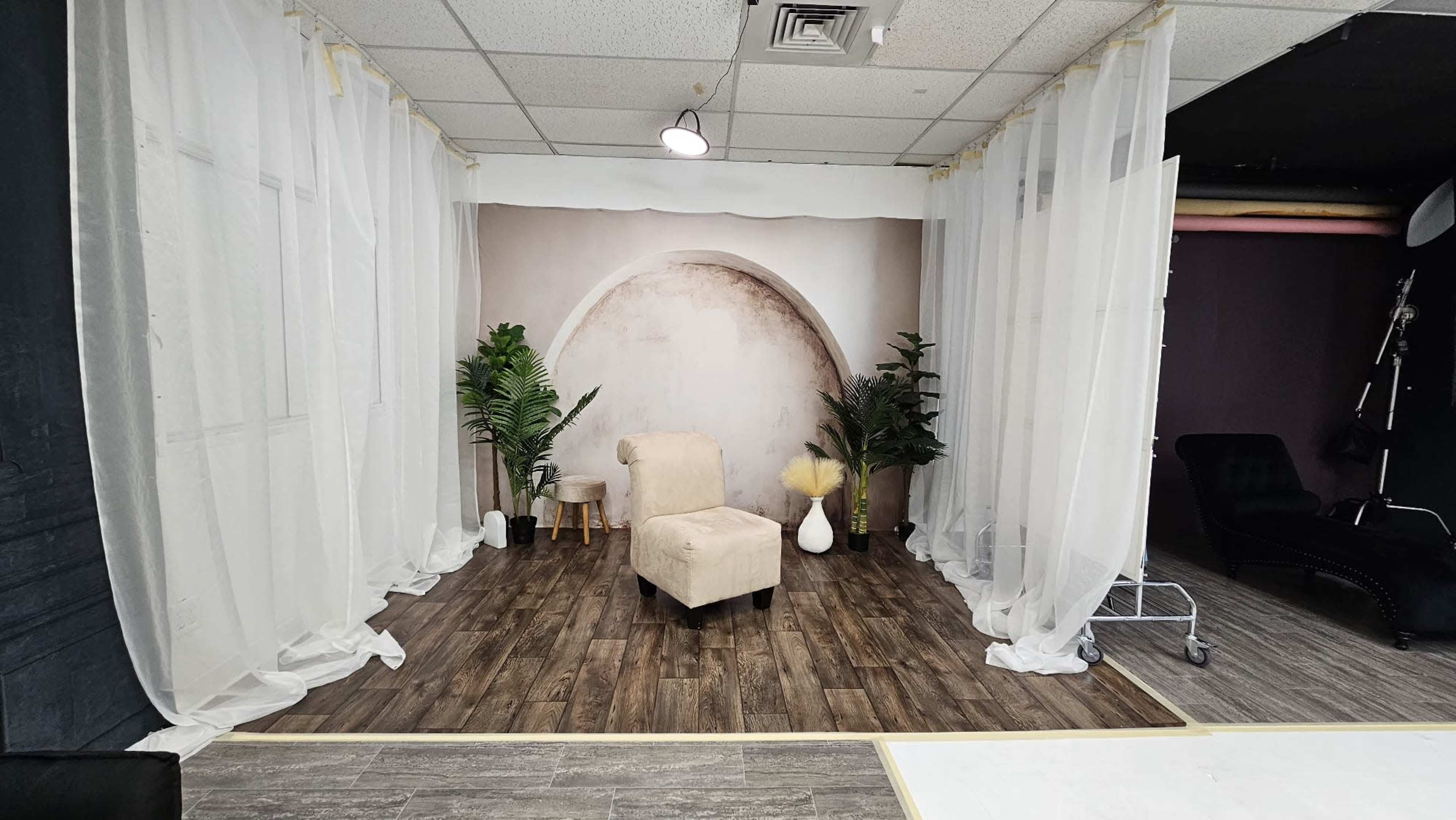 A studio setup with a light-colored armchair facing a backdrop of an arched design, surrounded by potted plants and soft drapery.