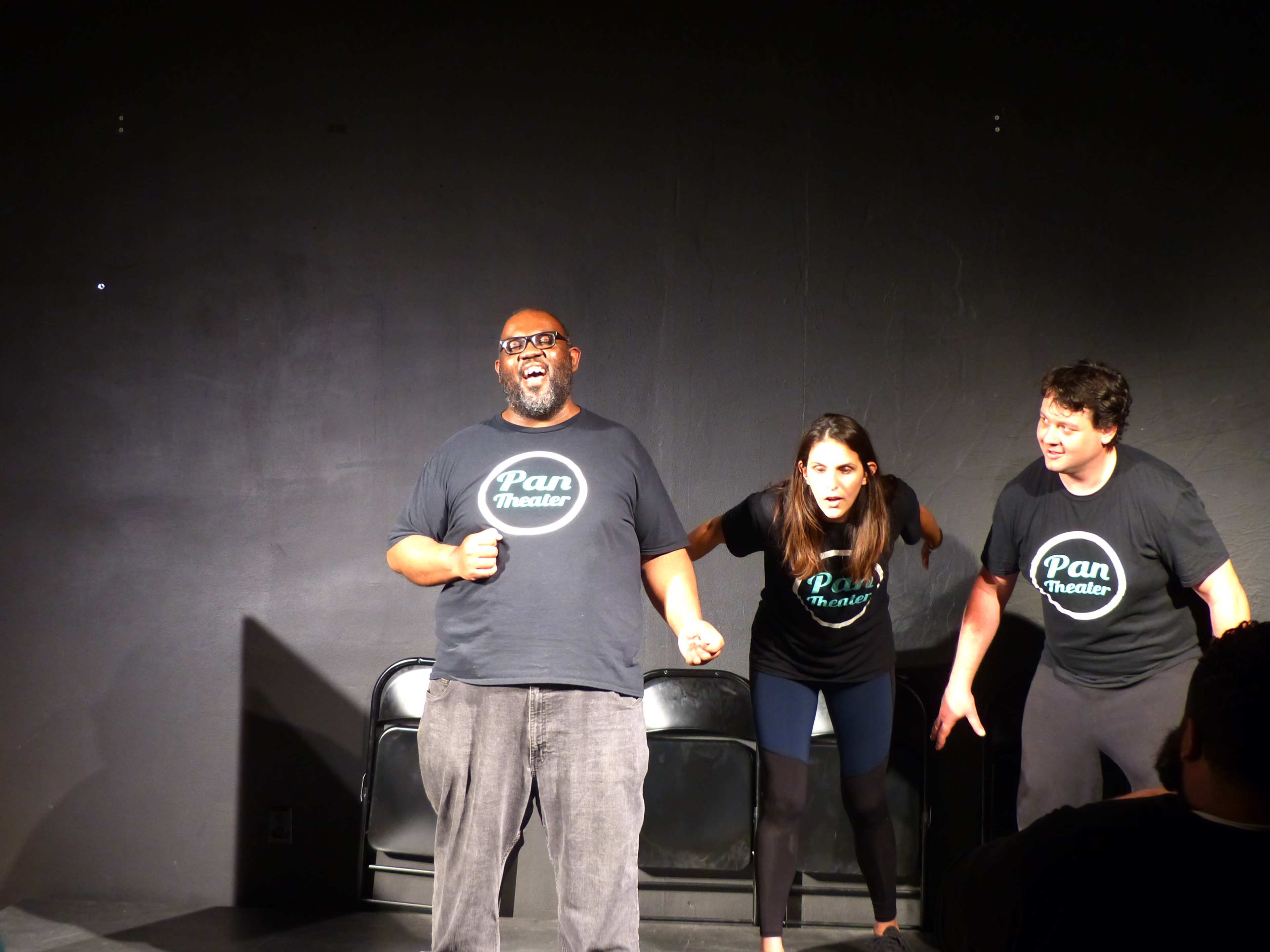 Three performers take the stage in black t-shirts with the "Pan Theater" logo, engaging in an energetic improv scene.