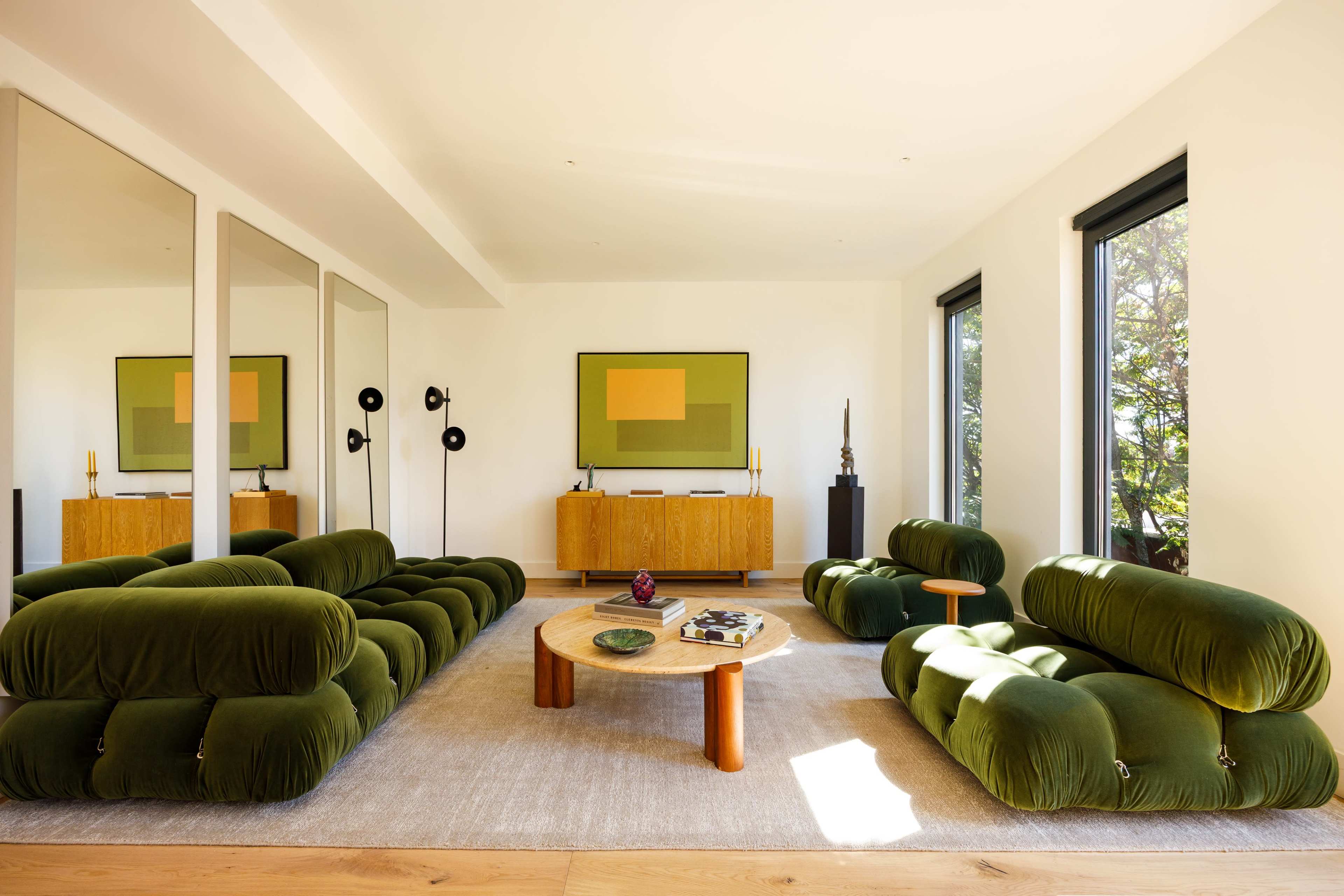 The image shows a modern living room with green upholstered sofas, a round wooden coffee table, and a large mirror reflecting natural light from large windows.